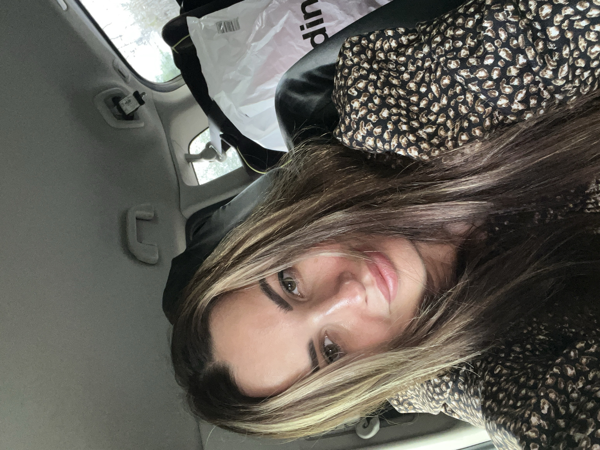 A woman with long, brown hair and streaks of blonde sits in the back seat of a car. She is wearing a dark-colored dress with a leopard-print pattern. Her eyes are open and she is looking at the camera with a slight smile. The background shows the interior of the car, including a white plastic bag with text on it, a seatbelt, and the roof of the car. The lighting appears to be natural, suggesting it is daytime.