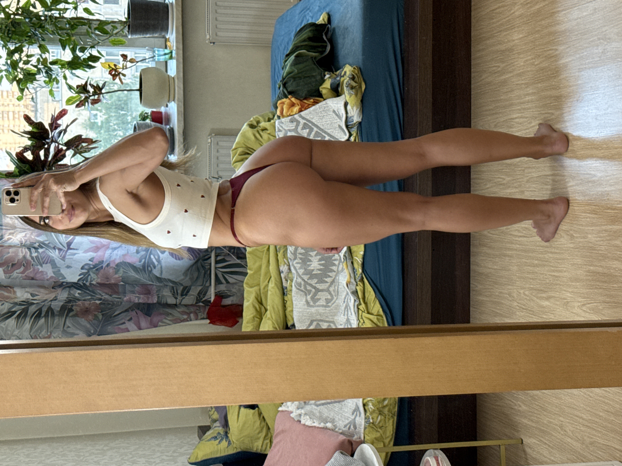 A woman stands in a bedroom, facing a mirror and taking a selfie. She is wearing a white cropped tank top with small red hearts and a maroon thong. Her hair is long and blonde, and she is wearing a bracelet on her right wrist. The room has a bed with green and yellow bedding, and a floral curtain behind her. There are plants on a windowsill, and radiators beneath the window. The floor is wood-effect laminate. The image appears to have been taken in a domestic setting, possibly an apartment, in an urban environment.