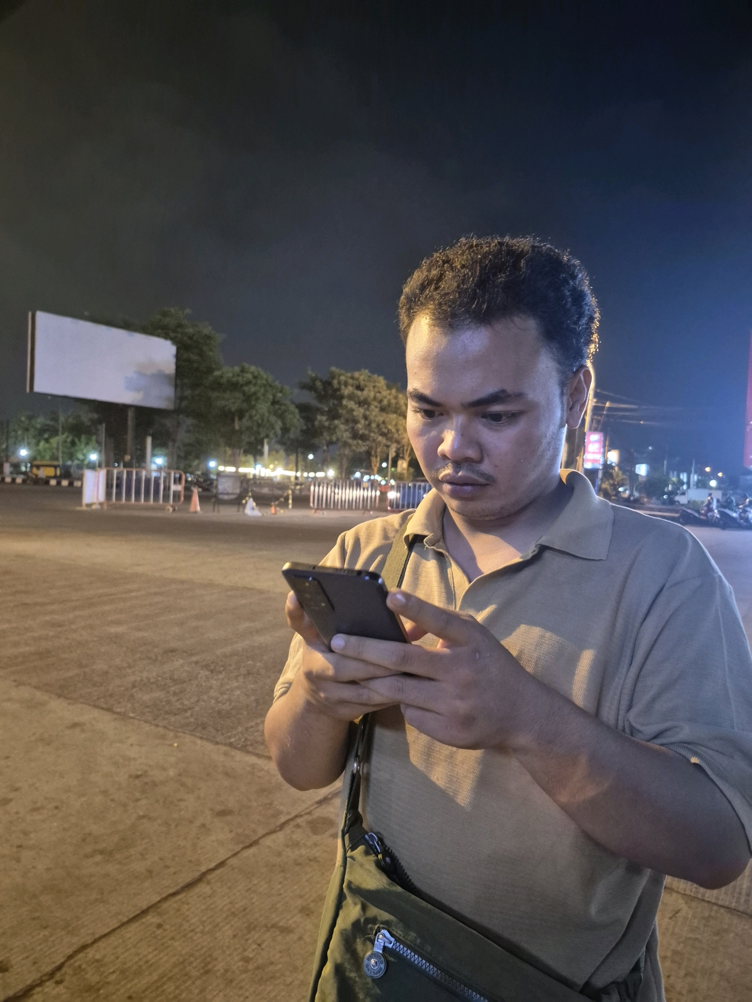 A medium shot captures a man standing at night, looking down at his black smartphone, which he holds with both hands. He is wearing a light brown collared shirt and a dark green messenger bag is slung across his body. The background is dimly lit, with a large blank billboard visible to the left. Several trees and streetlights are also present, suggesting an urban environment. The scene appears to be in the city of Surakarta, Indonesia, given the context provided.
