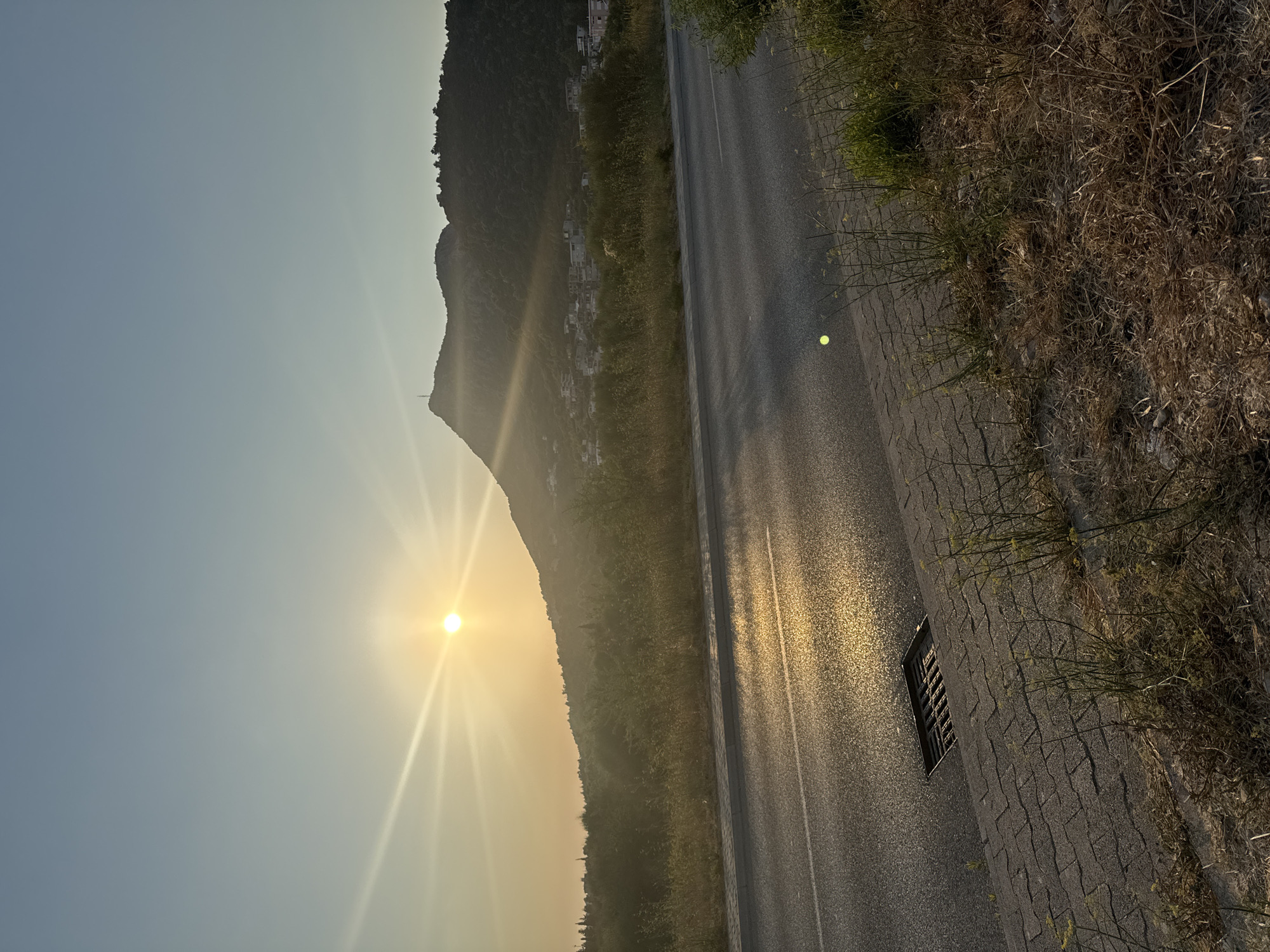 The image captures a sunset over a rural landscape near Sutomore, Montenegro. The sun is low in the sky, casting a warm glow and creating sunbeams. In the foreground, a paved road with a drainage grate is visible. To the right of the road, dry grass and some green vegetation are growing. Beyond the road, there is a hill covered in trees, with a few buildings dotting the hillside. The sky is clear and a pale blue.