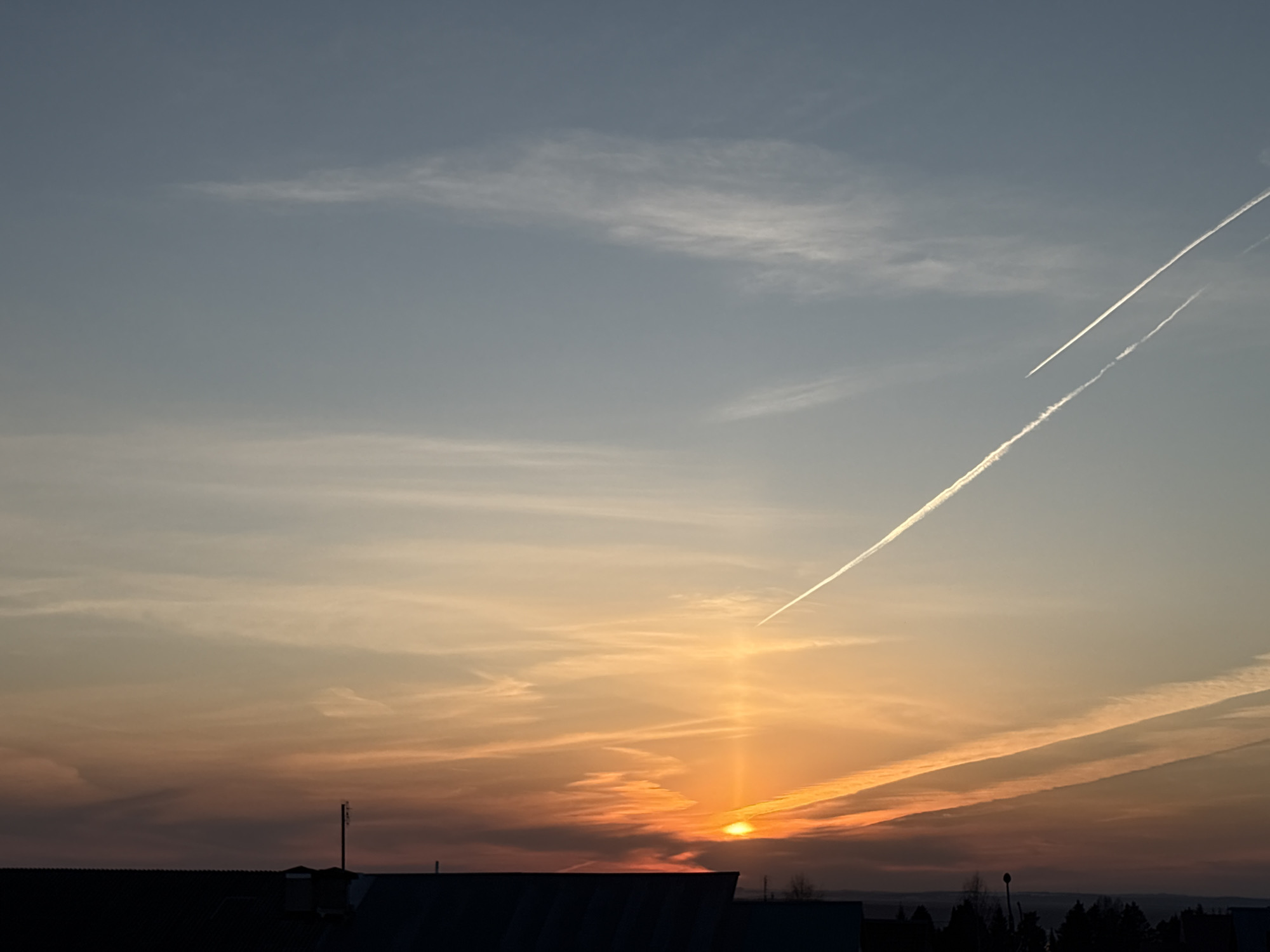 The image captures a sunset scene in Прикамский, Russia. The sun is near the horizon, emitting a bright orange glow, with a visible beam extending upwards into the sky. The sky is a mix of colors, transitioning from orange and red near the horizon to a lighter blue higher up. There are several contrails from airplanes crossing the sky. Below the horizon, there is the silhouetted skyline of buildings and trees. A single antenna stands out against the skyline.