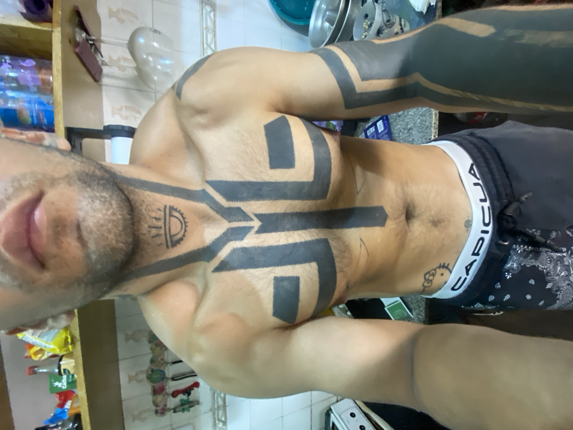 A close-up, indoor shot presents a man with extensive black geometric tattoos covering his arms, chest, and neck. A small tattoo of Hello Kitty can be seen near the waistband of his black patterned boxer shorts with a white elastic band labeled "CAPICUA." The background shows a typical domestic setting, with shelves displaying various food items and cleaning products, suggesting a kitchen or pantry area. The presence of white tiles on the wall and floor is also noticeable. The scene lacks any specific landmarks or context that would identify the location as Buenos Aires, Argentina.