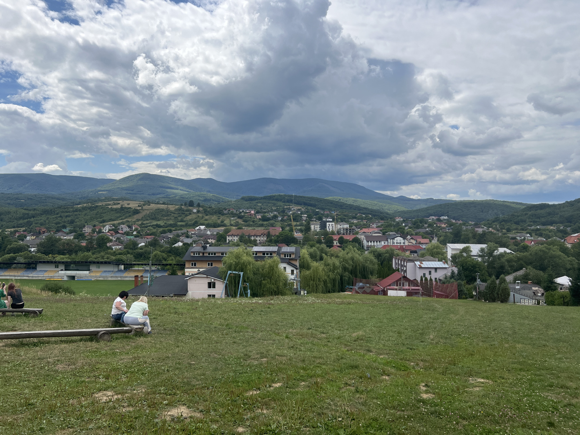 In Сонячне, Ukraine, on a grassy hillside overlooking a town, two people are sitting on a wooden bench. The person on the left is wearing a white t-shirt and jeans, and the person on the right is wearing a light blue t-shirt and white pants. They are facing away from the camera, towards the town and the mountains beyond. The town is nestled in a valley, with houses and buildings of various colors and sizes. Behind the town, rolling green hills rise up to meet a dramatic sky filled with fluffy, gray clouds. A stadium with blue and yellow seating is visible in the middle ground to the left of the frame. The overall scene is peaceful and serene, with a sense of natural beauty and quietude.