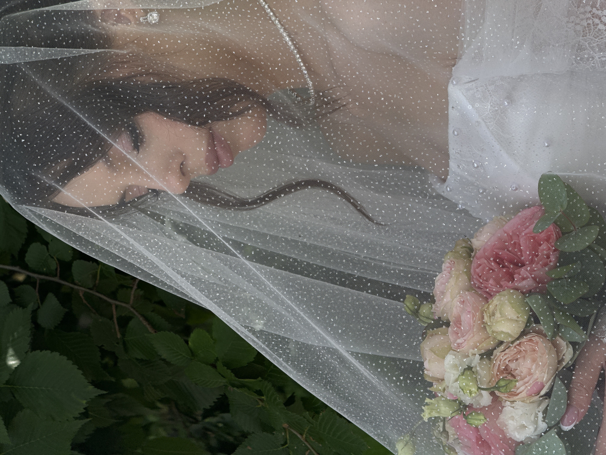 The image shows a close-up of a bride wearing a sparkling veil, holding a bouquet of pink and white roses. The background is a blur of dark green foliage, suggesting an outdoor setting. The bride's face is partially obscured by the veil, but her side profile is visible, with her eyes downcast. She is wearing a pearl necklace and diamond earrings. The bouquet contains various shades of pink and white roses, with some greenery. The veil is sheer and adorned with small, reflective dots. The lighting is soft, with the focus on the bride and her bouquet. The image appears to be taken in a natural environment, possibly a garden or park, typical of settings found in Votkinsk, Russia.