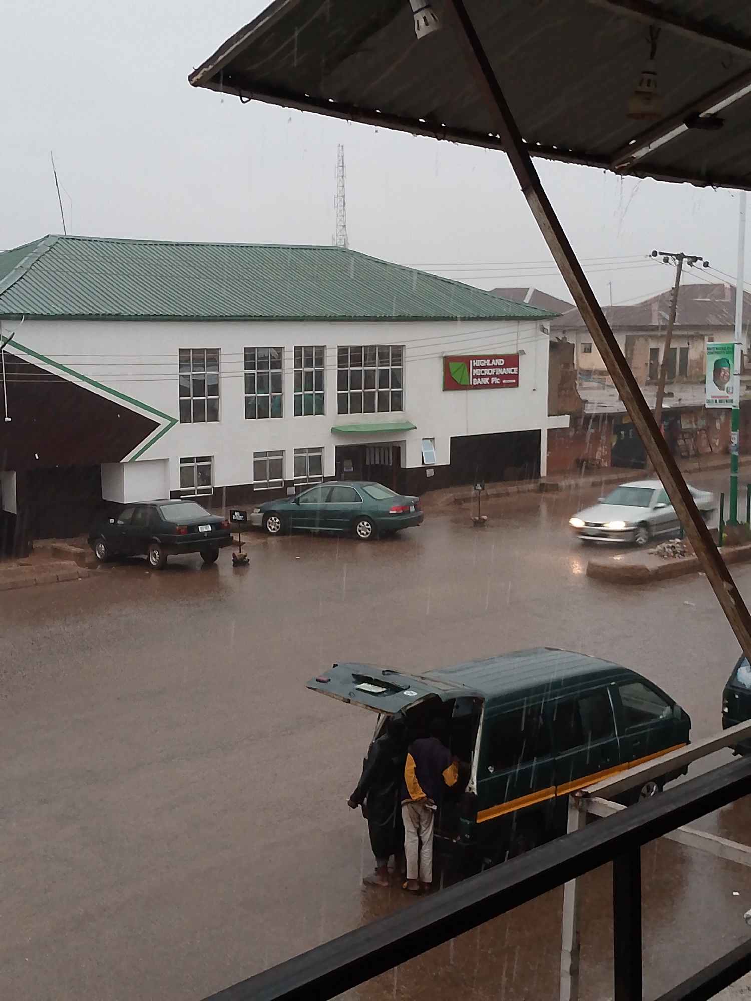 The image depicts a street scene in Bukuru, Nigeria, during heavy rain. In the foreground, two people are standing near the open rear of a dark green van with a yellow stripe. The street is flooded, reflecting the gray sky. A white building with a green roof and the sign "HIGHLAND MICROFINANCE BANK Plc" is visible in the background. Three parked cars are visible near the bank, as well as a fourth silver car in motion on the street. Utility poles and other buildings are also visible in the distance. The image is taken from an elevated position under a roofed structure.