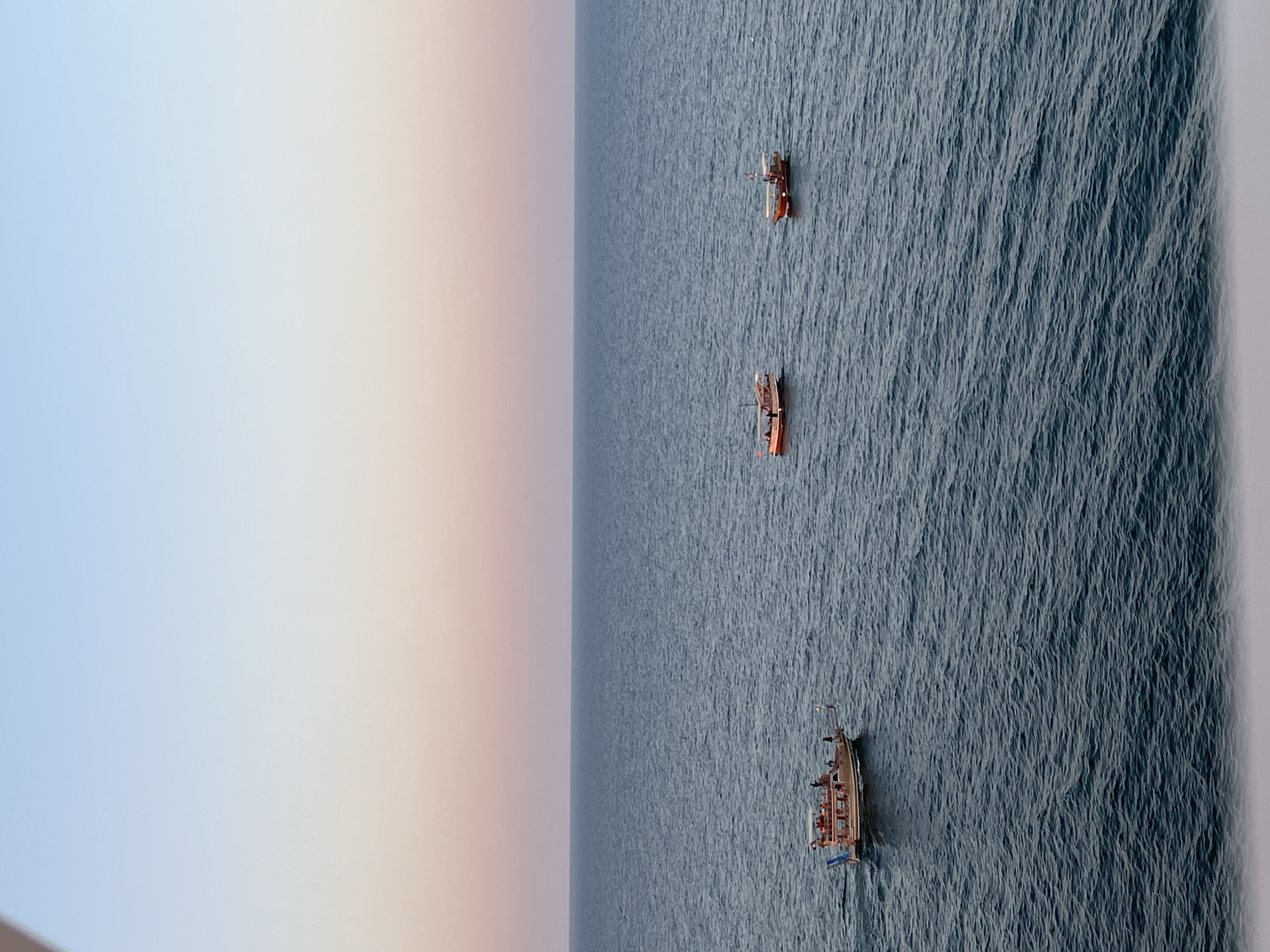 The image depicts a tranquil sea scene with three boats navigating the water under a gradient sky transitioning from pale blue to soft pink. The horizon line is clearly defined, separating the water from the sky. In the foreground, the ocean surface exhibits gentle ripples, indicating calm conditions.
One boat, located to the left, is a larger wooden vessel with multiple passengers visible on its deck. This boat is adorned with flags and appears to be a tour boat. Two smaller boats are positioned further to the right. Both of these boats are also wooden, with red accents on their structures. They appear to be moving at a similar pace as the larger boat. The overall setting suggests a coastal area, possibly near a city or harbor, during either sunrise or sunset due to the sky's coloration.
While the image is not explicitly set in Constanța, Romania, the general characteristics of a coastal city with maritime activity, such as boats on the sea, are consistent with locations like Constanța, which has a prominent port and coastline along the Black Sea. The presence of tour boats and smaller vessels is common in such areas.