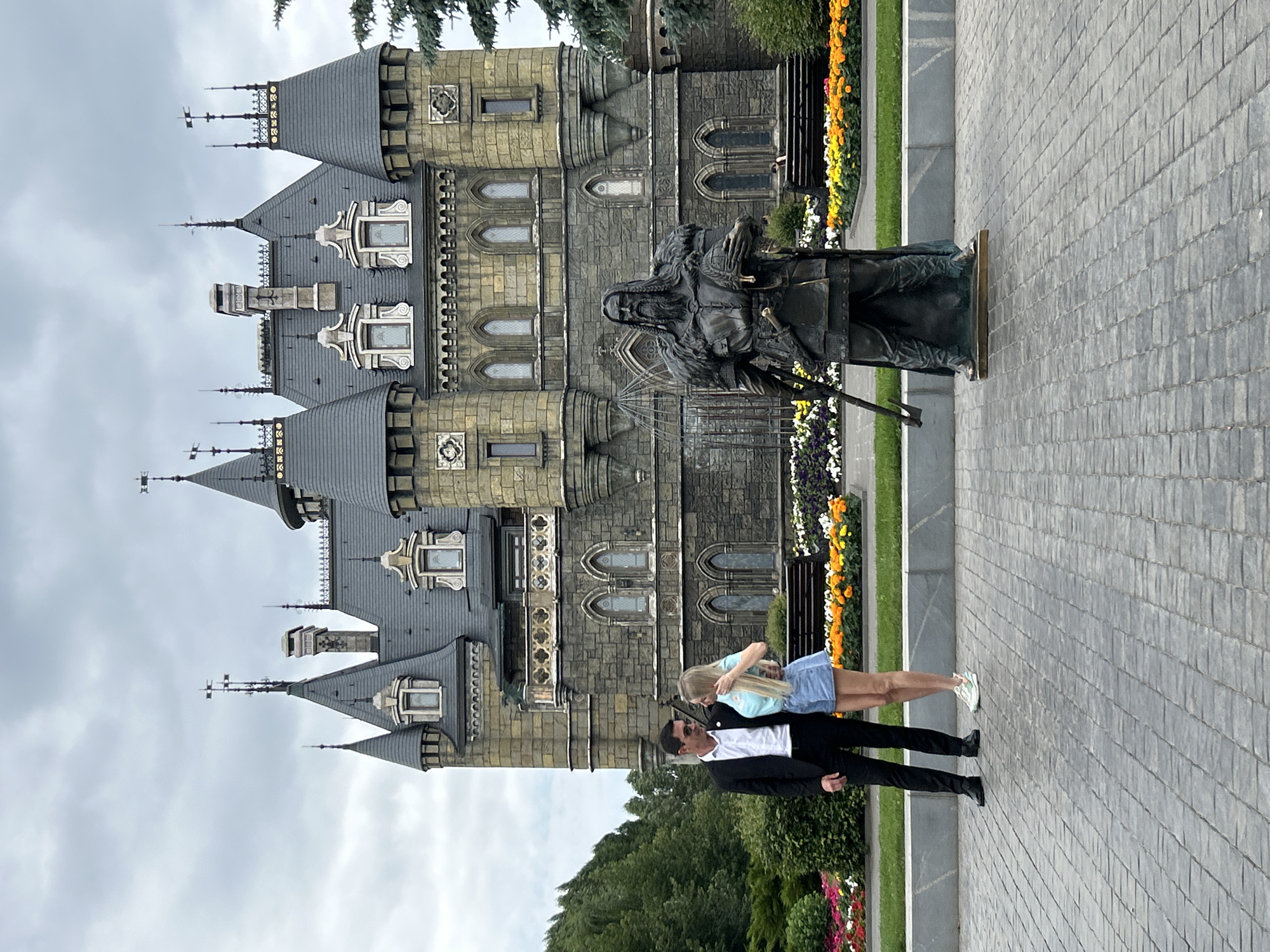 The image depicts a surreal, almost fairytale-like castle with multiple spires and towers, built with stone and dark grey roofing. In front of the castle, a bronze statue of a person with long hair and a beard stands stoically. Two people, a man and a woman, are visible in the foreground, facing the castle. The man is dressed in a black suit jacket over a white shirt and black pants, while the woman is wearing a light blue top and a denim skirt. They appear to be in a close embrace, with the woman's head resting on the man's shoulder.
The setting is outdoors, with manicured green lawns and flower beds featuring vibrant orange, yellow, and white flowers lining the path in front of the castle. The ground in the foreground is paved with grey cobblestones. The sky is overcast and cloudy, suggesting a temperate day. The architecture of the castle, with its gothic-inspired windows and intricate details, evokes a sense of fantasy or historical drama. The overall mood is one of romance and contemplation, set against a grand and somewhat imposing backdrop. The location is identified as Khryashchevka, Russia. No visible text is present in the image.