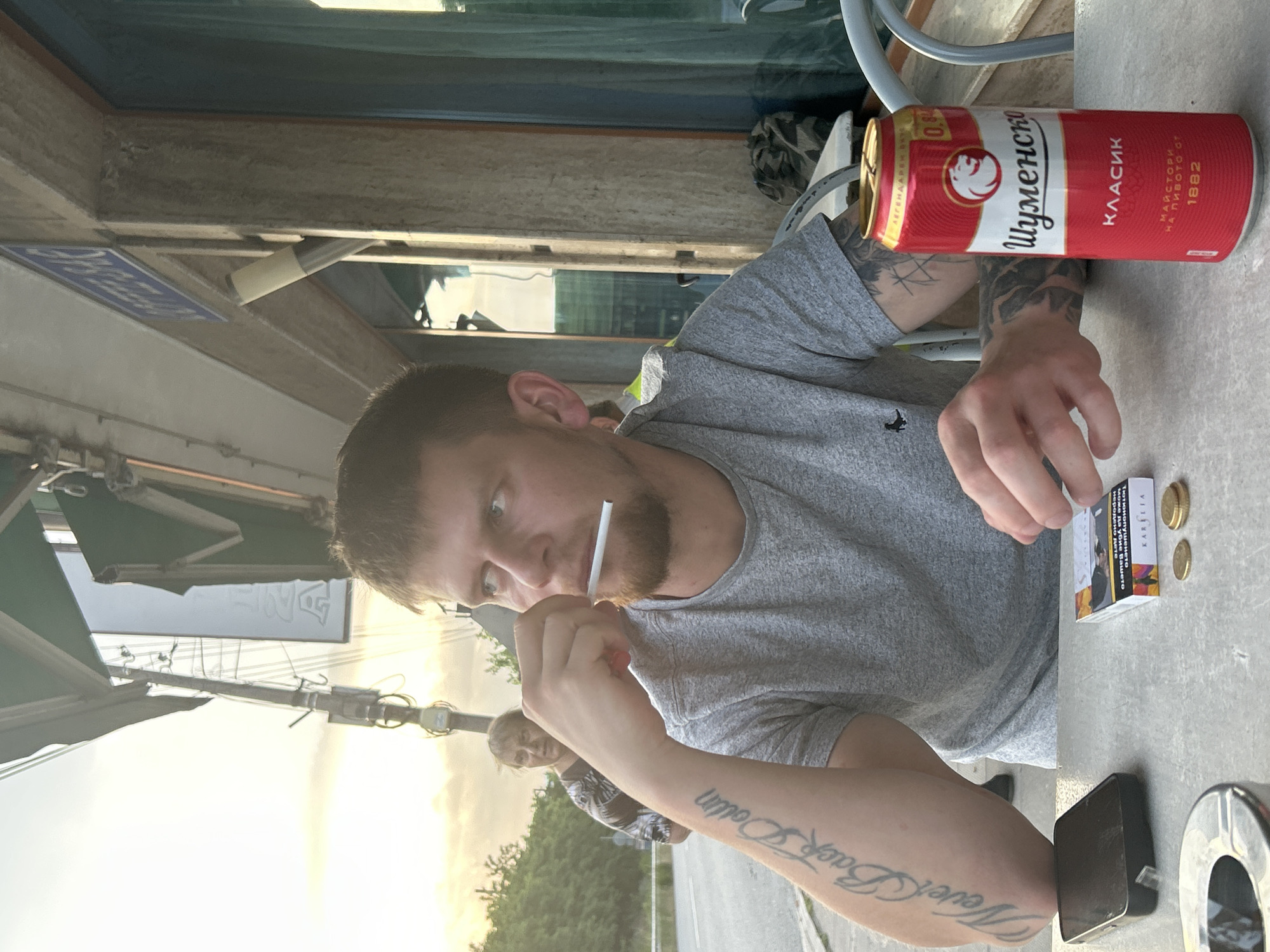 The image depicts an outdoor patio scene at dusk in the city of Unknown, Unknown. A young adult male, with a beard and short brown hair, is seated at a light-colored table, wearing a gray t-shirt with a small dark logo on the left chest. He holds an unlit white cigarette or filter between his lips, gazing intently towards his right. Tattoos are visible on both forearms; the left forearm displays "Never Back Down" in script. On the table are a red and white aluminum can of 'Шуменско КЛАСИК' (Shumensko Classic) beer, marked 0.0% alcohol, a pack of 'KARELIA' cigarettes, two gold-colored coins, a black smartphone lying screen-down, and a partially visible metal ashtray. In the background, across a street with a curb, an older adult female with gray hair and a patterned top stands, looking towards the man. The sky above exhibits warm, golden-hour lighting. Architectural elements include a green awning, a light fixture, and a blue sign with white text mounted on the adjacent building wall.