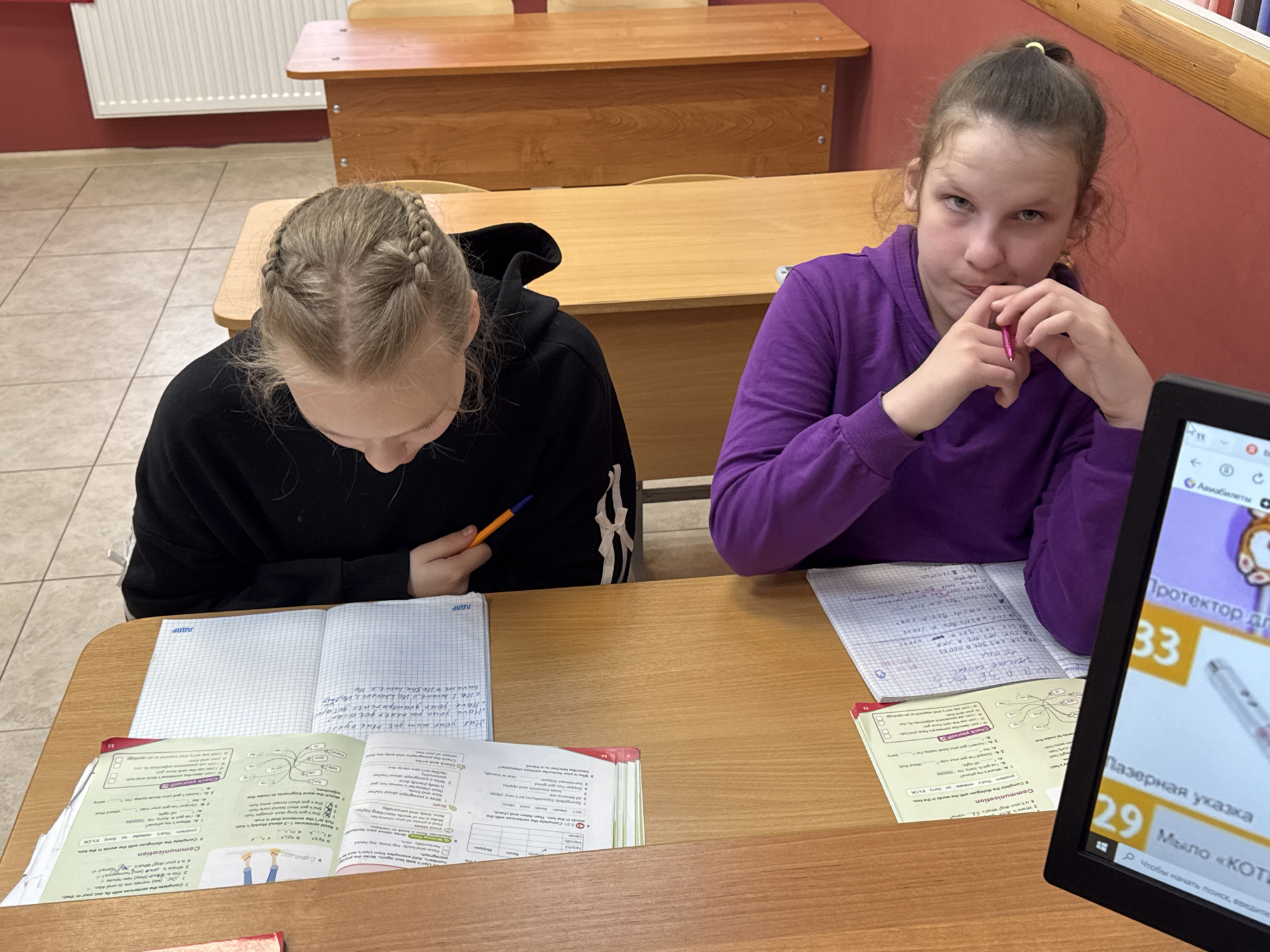 Two young girls, likely students, are depicted studying at individual wooden desks in what appears to be a classroom in Perm, Russia. The room features a tiled floor and a red wall with a white radiator and additional wooden desks in the background, characteristic of an educational institution.
The girl on the left, with light-colored hair styled in two French braids, is seen from above, deeply focused on her open notebook and English textbook. She holds an orange pen, ready to write. Her notebook contains handwritten English questions such as "Have your mum got blue eyes?" and "Have your parents got dark hair," along with the Russian acronym "ЛДПР." Her textbook, open to pages 8 and 55, displays English language exercises, including "Communication" tasks, matching word fragments, and dialogue completion.
To her right, another girl with darker hair, tied in a ponytail with a yellow band, wears a purple long-sleeved top. She looks directly towards the viewer with a thoughtful or slightly intense expression, holding a pink pen near her mouth. Her desk also holds an open notebook and textbook, suggesting she is engaged in similar academic work.
A black computer monitor is partially visible on the right side of the frame, displaying various items. The screen shows Russian text including "Авиабилеты" (Air tickets), "Протектор дл" (Protector for), "Лазерная указка" (Laser pointer), and "Мыло «Кот" (Cat Soap), accompanied by numbers "33" and "29," and a prompt "Чтобы начать поиск, введите" (To start the search, enter). These details indicate the potential use of online resources or educational software during their study session. The indoor lighting does not provide specific clues about the time of day.