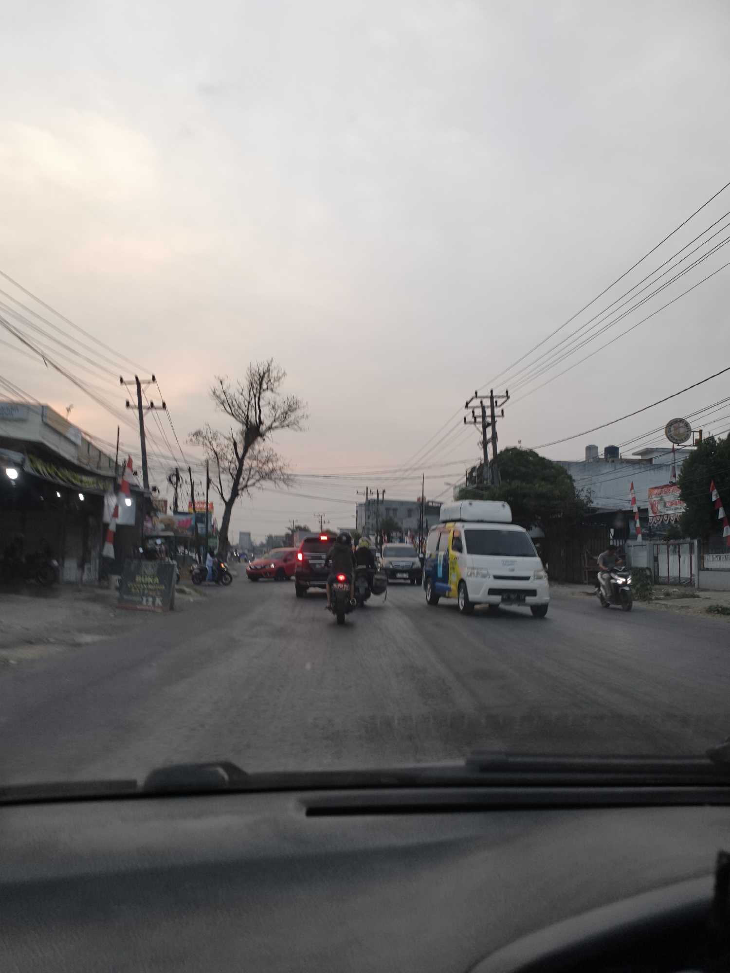 The image captures a street scene in Kemala Raja, Indonesia, during what appears to be dusk. The sky is overcast with a pale orange glow, suggesting the sun is low on the horizon. The road is wet, indicating recent rain. Various vehicles are present, including cars, motorcycles, and a white van. Several people are visible riding motorcycles. The roadside features buildings, some with awnings and lights, and utility poles with numerous overhead wires. A leafless tree stands prominently near the center of the frame. The perspective is from inside a vehicle, looking out through the windshield, parts of which are visible in the foreground.