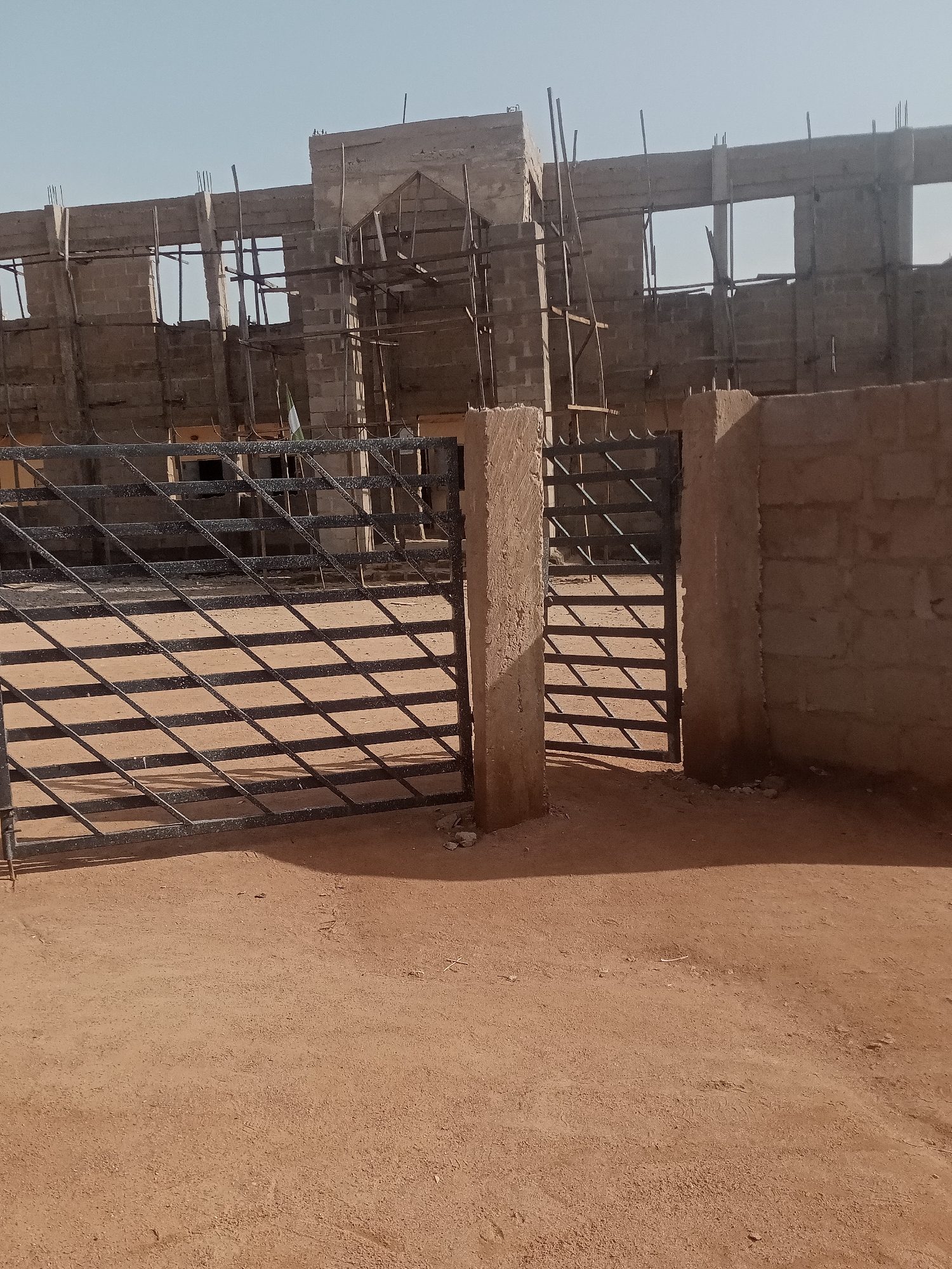 This image depicts a construction site in Jalingo, Nigeria. In the foreground, a black metal gate with a diamond pattern is visible, casting shadows on the dusty ground. The gate is partially open, revealing more of the construction area. To the right of the gate, a rough, concrete pillar stands, and beyond it, a wall constructed from concrete blocks extends horizontally. Behind the gate and wall, a multi-story building is under construction. The building appears to be made of concrete blocks and has exposed rebar and wooden scaffolding. Several windows are visible, some are open spaces, and others appear to be framed. A flag, possibly the Nigerian flag with its green and white stripes, is attached to the scaffolding of the building. The sky is clear and pale blue. The ground is predominantly reddish-brown soil, indicative of arid or semi-arid conditions.
