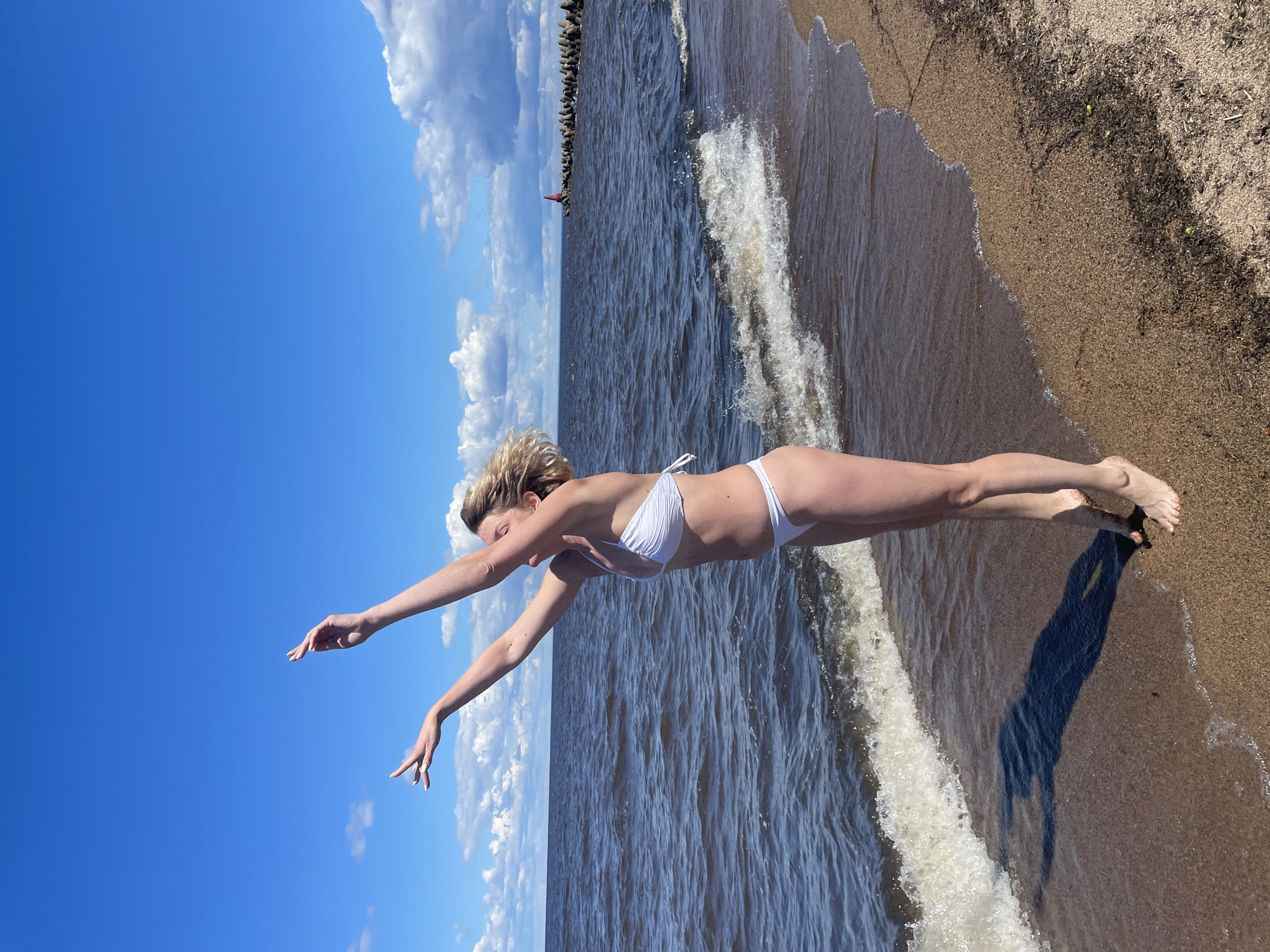 A woman in a white bikini stands on a sandy beach, her arms raised to the sky. She is barefoot, and her shadow is cast on the wet sand. The sea is choppy with waves crashing on the shore. In the distance, a breakwater extends into the water, topped with a red buoy. The sky is blue with scattered white clouds. The scene is set in Belozersk, Russia.