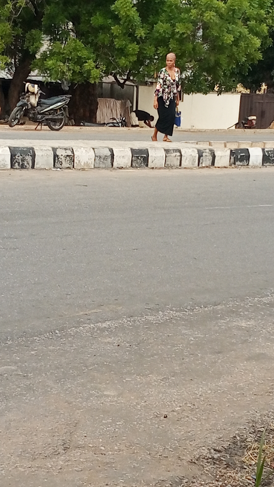 A woman walks to the right on a sidewalk. She is wearing a black skirt and a floral top. She has a blue bag on her left arm. Behind her on the sidewalk, a person is bent over. There is a motorcycle parked to the left. A black and white curb divides the sidewalk from a wide paved road. The foreground is the road and a dirt shoulder. In the background, a green tree line is visible. The location is in the city of Adankolo, Nigeria.