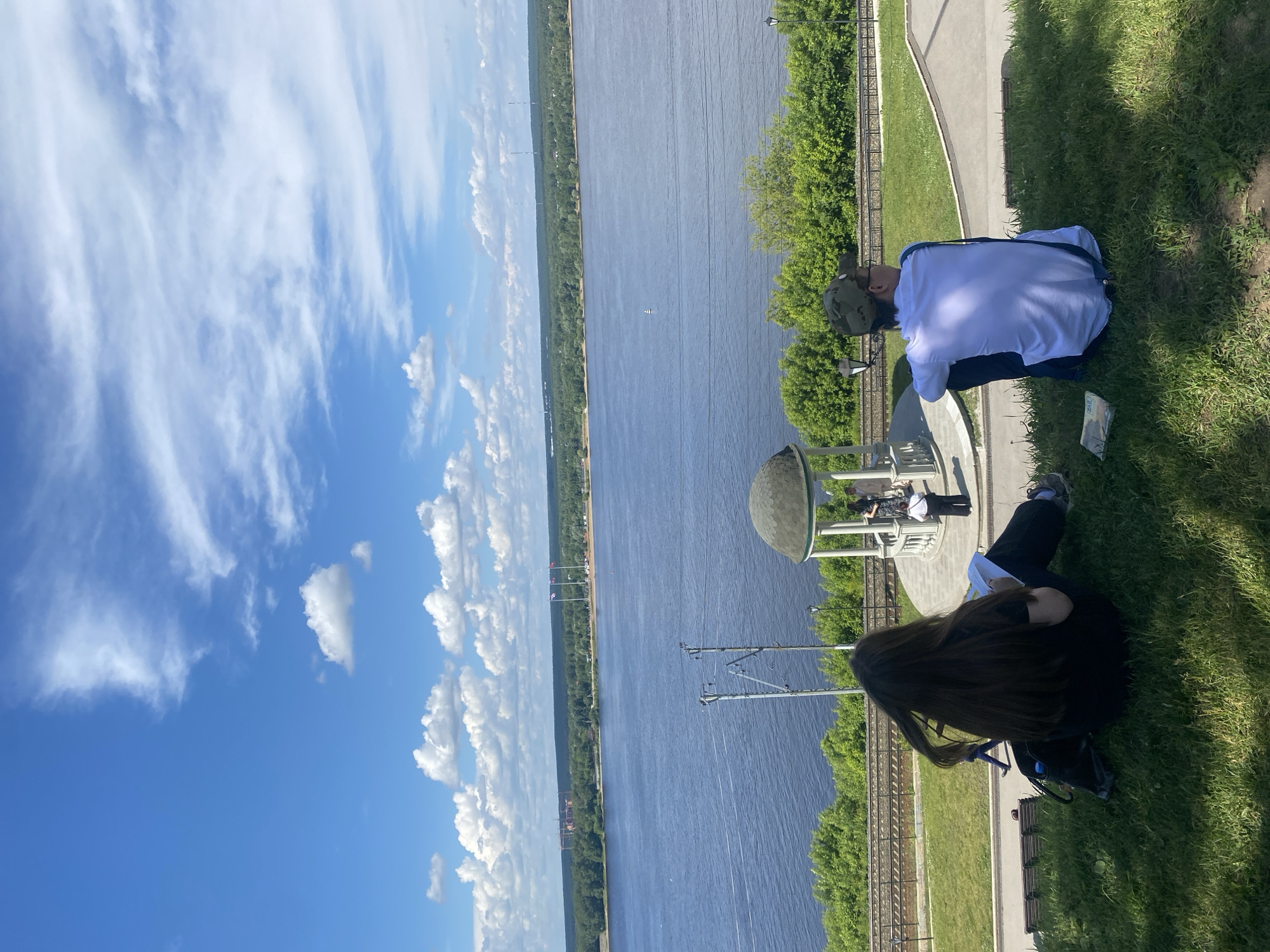 The image shows a scenic view of the Kama River in Perm, Russia. Two individuals are seated on a grassy hill overlooking the river. One person, with long dark hair, is facing away from the camera, engrossed in a book or notebook. The other person, wearing a baseball cap and a white shirt with blue suspenders, is also looking out at the river. In the middle ground, a gazebo stands near the riverbank, and a few other people are visible near it. Train tracks run along the embankment below the hill, and lush green trees line the river. The sky is blue with scattered white clouds. The overall atmosphere is peaceful and relaxed, suggesting a pleasant day outdoors.
