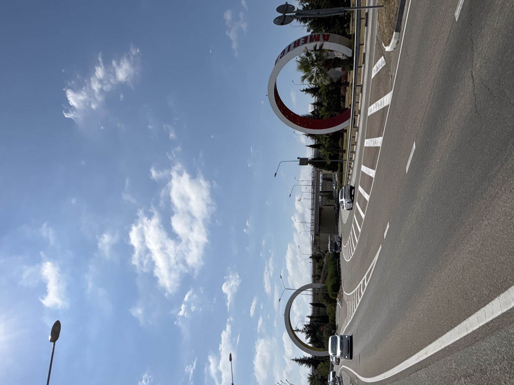 The image shows a daytime view of a modern road with white lane markings curving through a landscaped area. Two large, circular arch structures are visible; one on the left is white and appears to be an entrance arch, while the one on the right is white with red interior and has the word "AMERI" visible on its side. Several vehicles, including cars and a bus, are driving on the road. Green trees and palm trees are scattered throughout the scene. In the background, an elevated road or bridge structure is visible, and the sky is blue with scattered white clouds. The scene is located in Sochi, Russia, possibly near an airport or a major transportation hub, given the scale of the structures and road infrastructure.