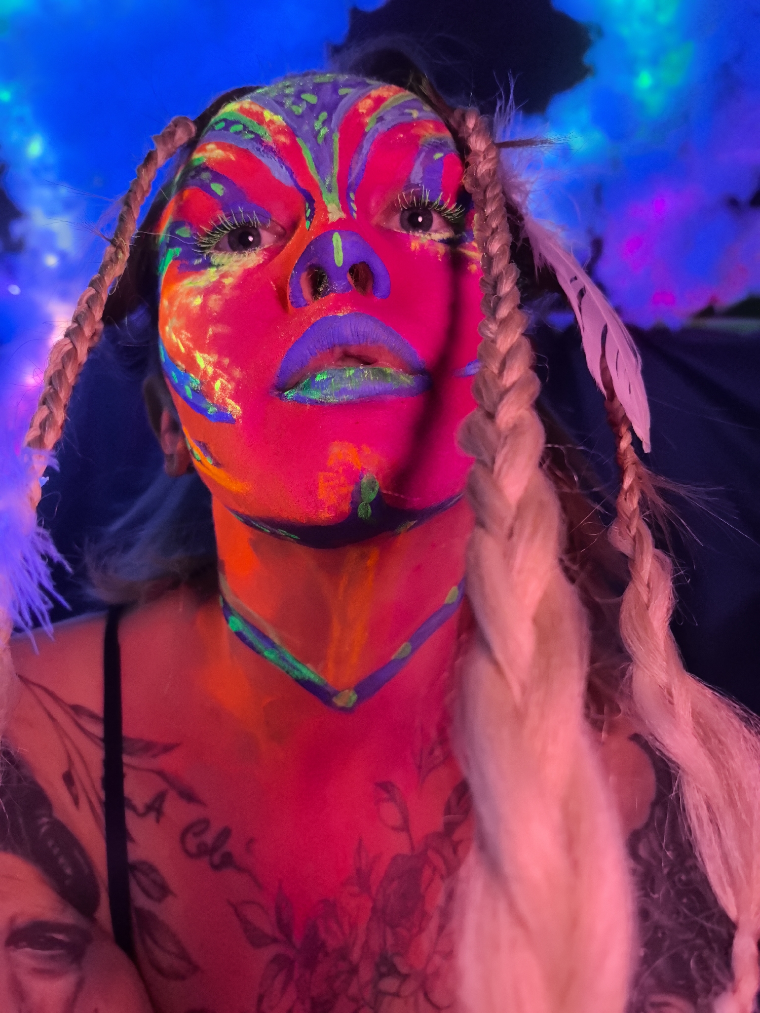 The image presents a striking, close-up portrait of a person with elaborate, neon-glowing face and body paint, captured under blacklight in what appears to be an indoor setting in Noviy, Russia. The subject's face is heavily adorned with vibrant, fluorescent pink/red paint, overlaid with intricate patterns in neon blue, purple, green, and orange. The eyes are open and gaze directly at the viewer with an intense expression, and the eyelashes are highlighted with luminous green. The lips are painted a vivid purple with a splash of green.
The individual has light-colored hair styled into long braids, one visible over each shoulder, incorporating white feathers. A dark clothing strap is visible on the left shoulder. On the left side of the chest and shoulder, several tattoos are visible, including cursive text that resembles "A Cole" or "A Cola," alongside floral or organic designs. The background is a vibrant blur of glowing blue and purple, suggesting a smoky or cloudy effect under UV light, creating a surreal and energetic atmosphere. The strong blacklight illumination makes the fluorescent paints on the skin glow intensely, highlighting the artistic makeup and creating a captivating visual. The scene suggests an artistic performance, a themed event, or a creative photoshoot, likely taking place in a dark environment at night.