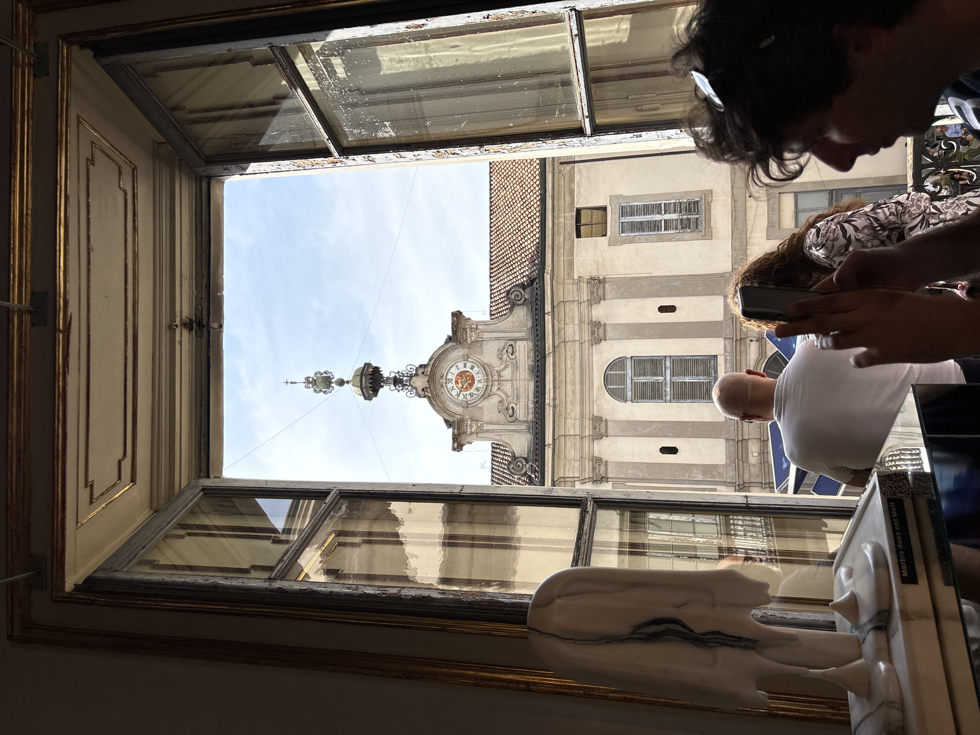 A slightly low-angle indoor shot through an old window looking out onto a building. The window is open and the light outdoors suggests it's daytime. There are several panes of glass in the window, some reflecting the interior. The window frame is ornate.
Outside, the primary focus is a building with a clock tower. The clock is visible and has Roman numerals. The building appears old and ornate.
In the foreground, near the window, there are people. One person is looking at a mobile phone. There is a bald person in a white shirt to the left. There is a sculpture or object made of white marble.
The scene is in Milan, Italy, specifically focused on the tower of the Palazzo della Ragione.