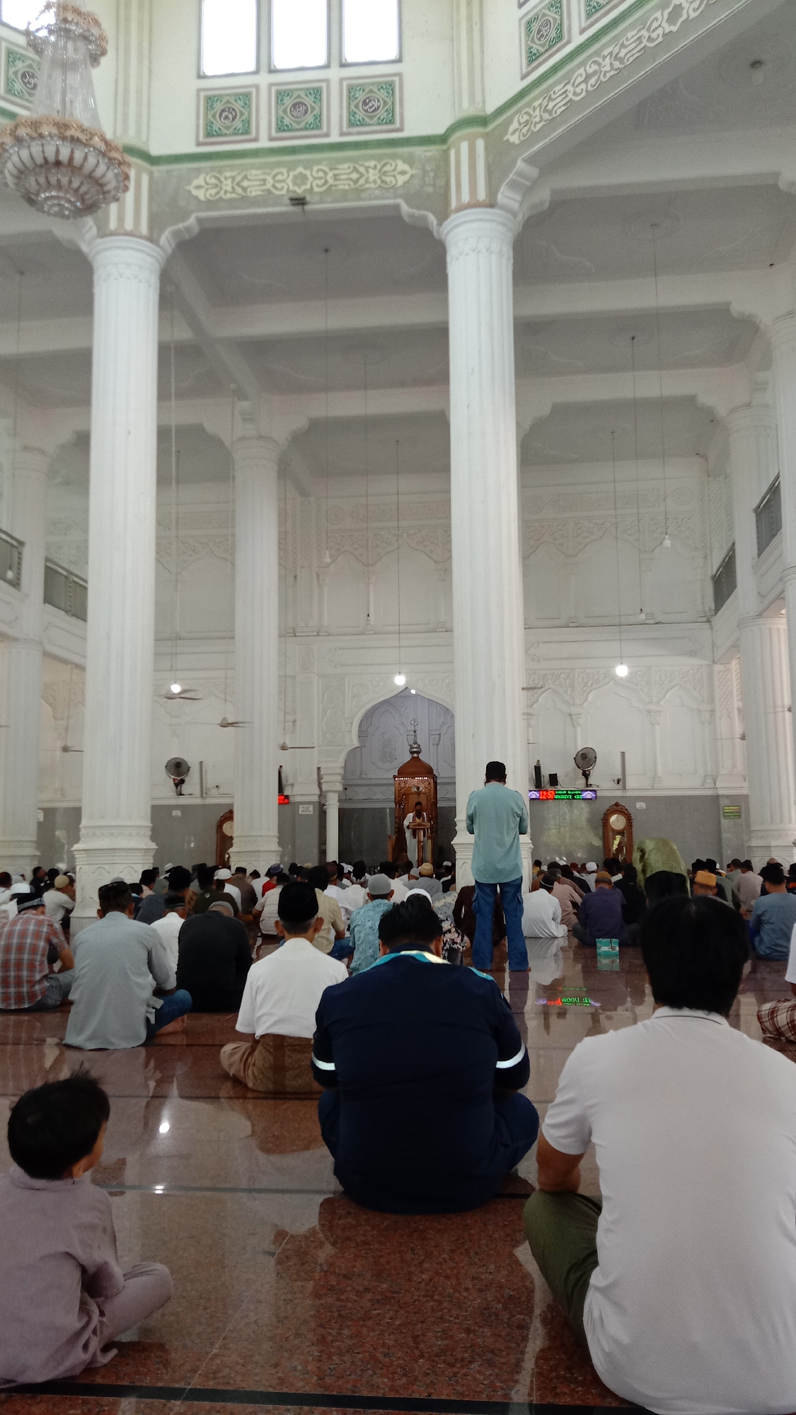This image depicts the interior of a spacious mosque in Lhokseumawe, Indonesia, during what appears to be a religious gathering or prayer session. The perspective is from the back of the prayer hall, looking towards the mihrab and minbar.
Numerous Muslim men and boys are seated on a polished, reddish-brown tiled floor, facing the front of the mosque. Most have their backs to the camera, indicating their focus on the proceedings. A few individuals are standing in the mid-ground. At the far end, a wooden minbar (pulpit) is visible, where a person is standing, likely delivering a sermon or leading prayer.
The mosque's architecture is grand, featuring several tall, white, fluted columns that support a high ceiling. The ceiling and upper walls are intricately decorated with white stucco patterns, arched sections, and green accents. A large, ornate chandelier hangs from the ceiling in the upper left. Wall-mounted fans are visible on the side walls, suggesting air circulation. On the right wall, a digital clock display shows "12:56," likely indicating the time, with some smaller, illegible text below it. Further up the walls, several square plaques with what appears to be Arabic calligraphy add to the decorative elements. The overall scene suggests a calm and reverent atmosphere during a communal religious event.