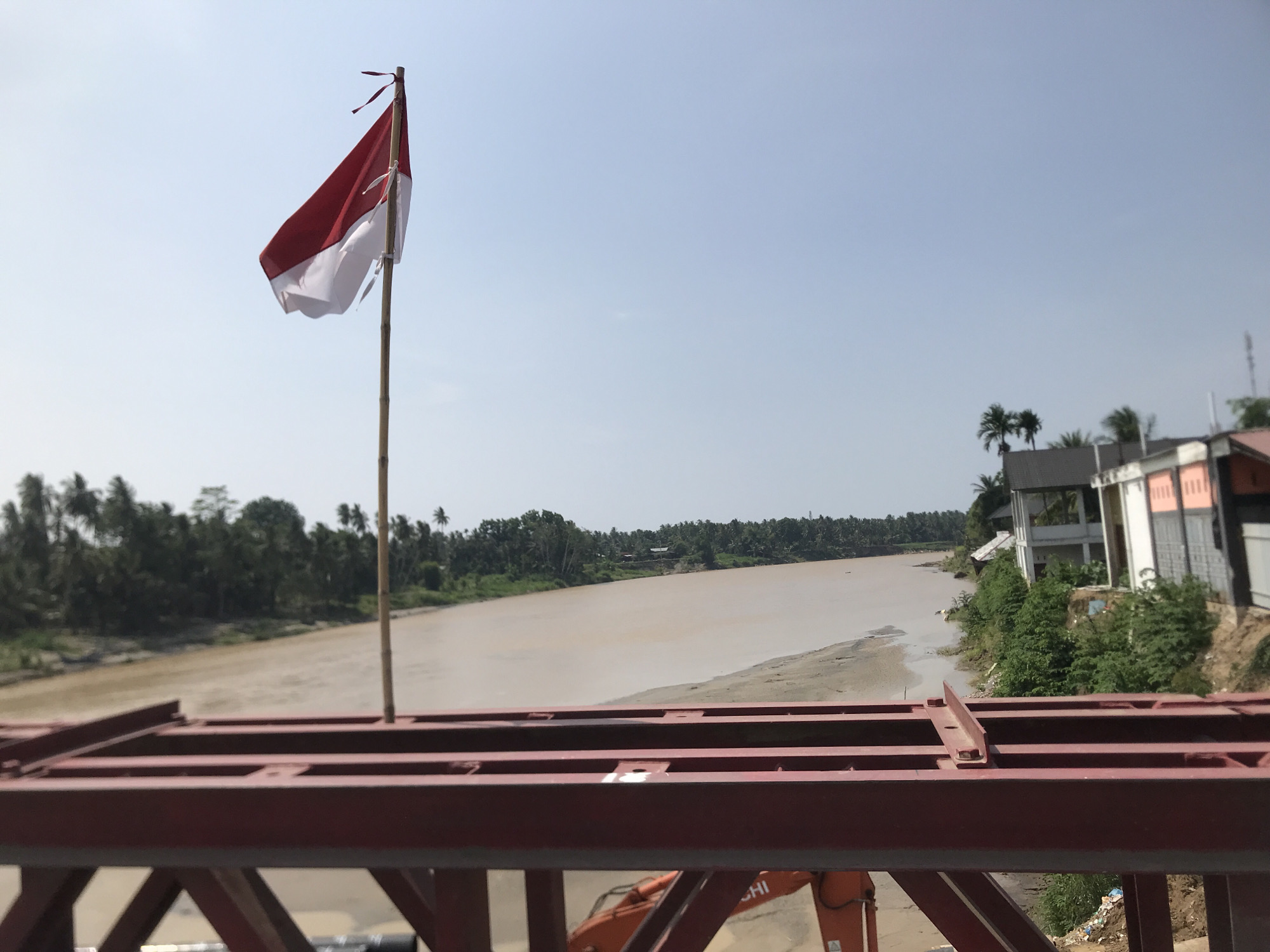 A horizontal, eye-level shot reveals a river in Blang Mee, Indonesia. The river is murky brown and calm. The shot is taken from a red, unfinished metal bridge with girders and beams. In the foreground, the bridge's metal frame is prominently displayed.
To the left and right of the river are shorelines with trees and vegetation. On the right side of the river, near the shoreline, are several buildings with varying designs and colors. An Indonesian flag is on the left with red over white.
In the background, the sky is a clear, pale blue with no clouds visible. An orange excavator can be seen on the right.