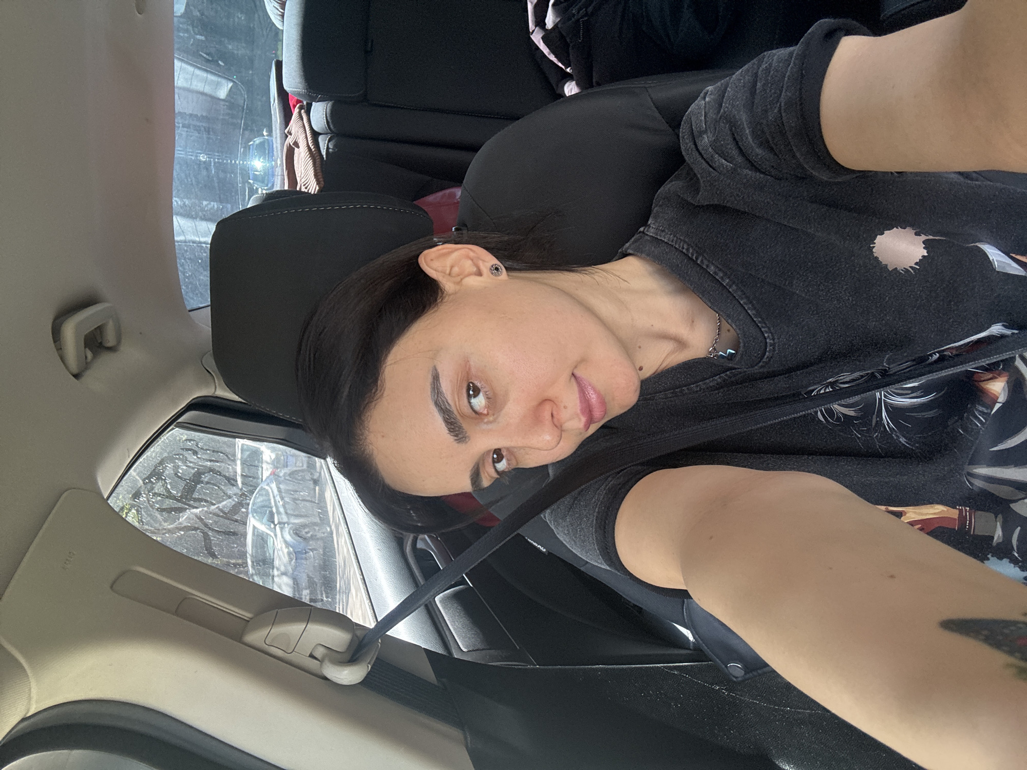 A woman is sitting in the back seat of a car, wearing a seatbelt and facing the camera. She has dark hair and is wearing a dark-colored shirt or jacket. The car's window is visible in the background, showing trees. The location is Tbilisi, Georgia.