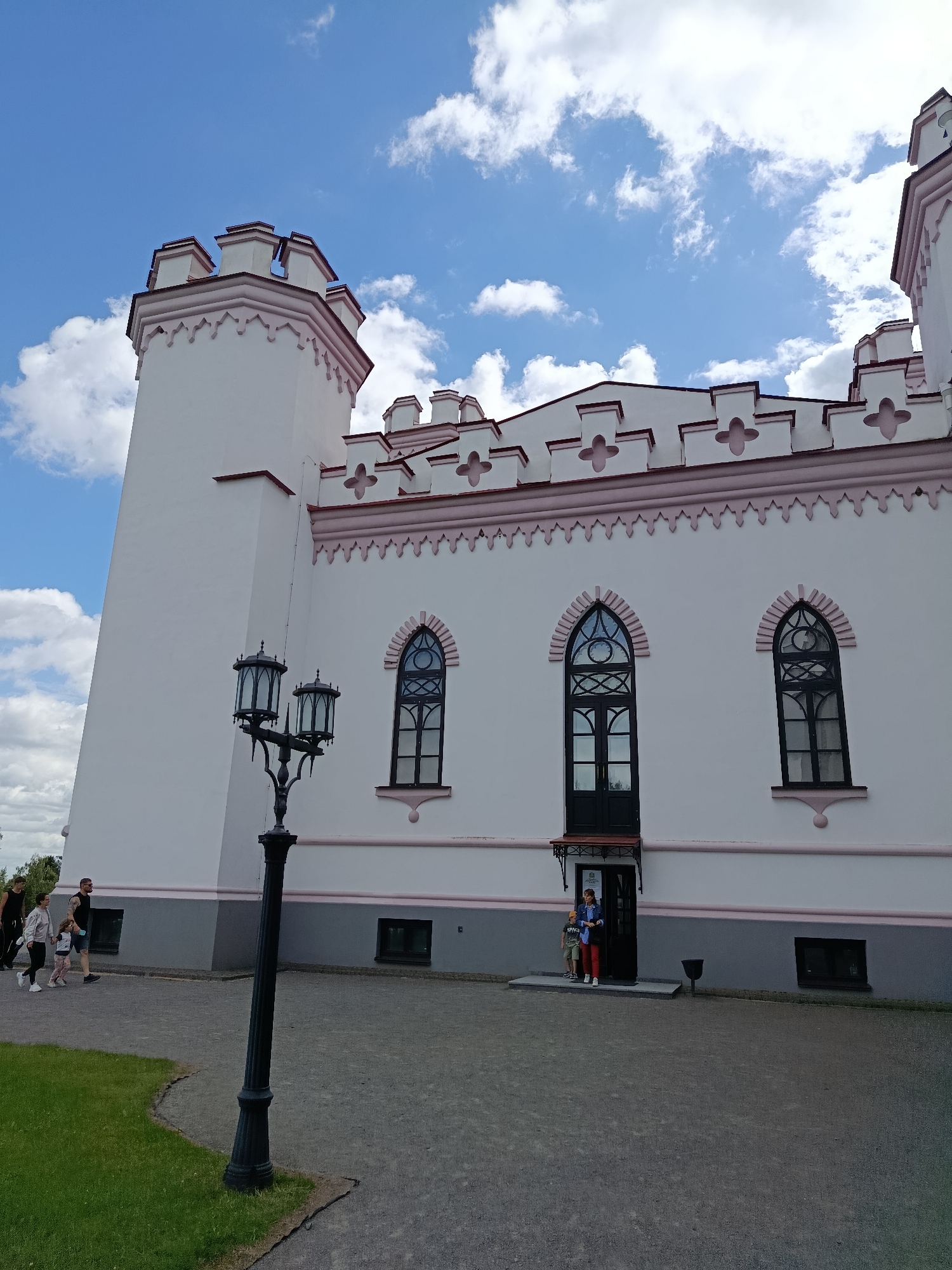 This is a photograph of the Kosava Palace in Kosava, Belarus, under a partly cloudy blue sky. The palace is a white structure with pink decorative trim along its crenellated roofline and around its arched windows. A prominent tower is visible on the left side of the building. In front of the palace, a black, ornate lamppost stands tall, its three lanterns facing outward. Several people are visible on the grounds. On the left, a group of four individuals, including two adults and two children, walk past the building. Near the entrance to the palace, a woman and a young boy stand on the steps. The grounds are paved with gravel, with a patch of green grass to the left.