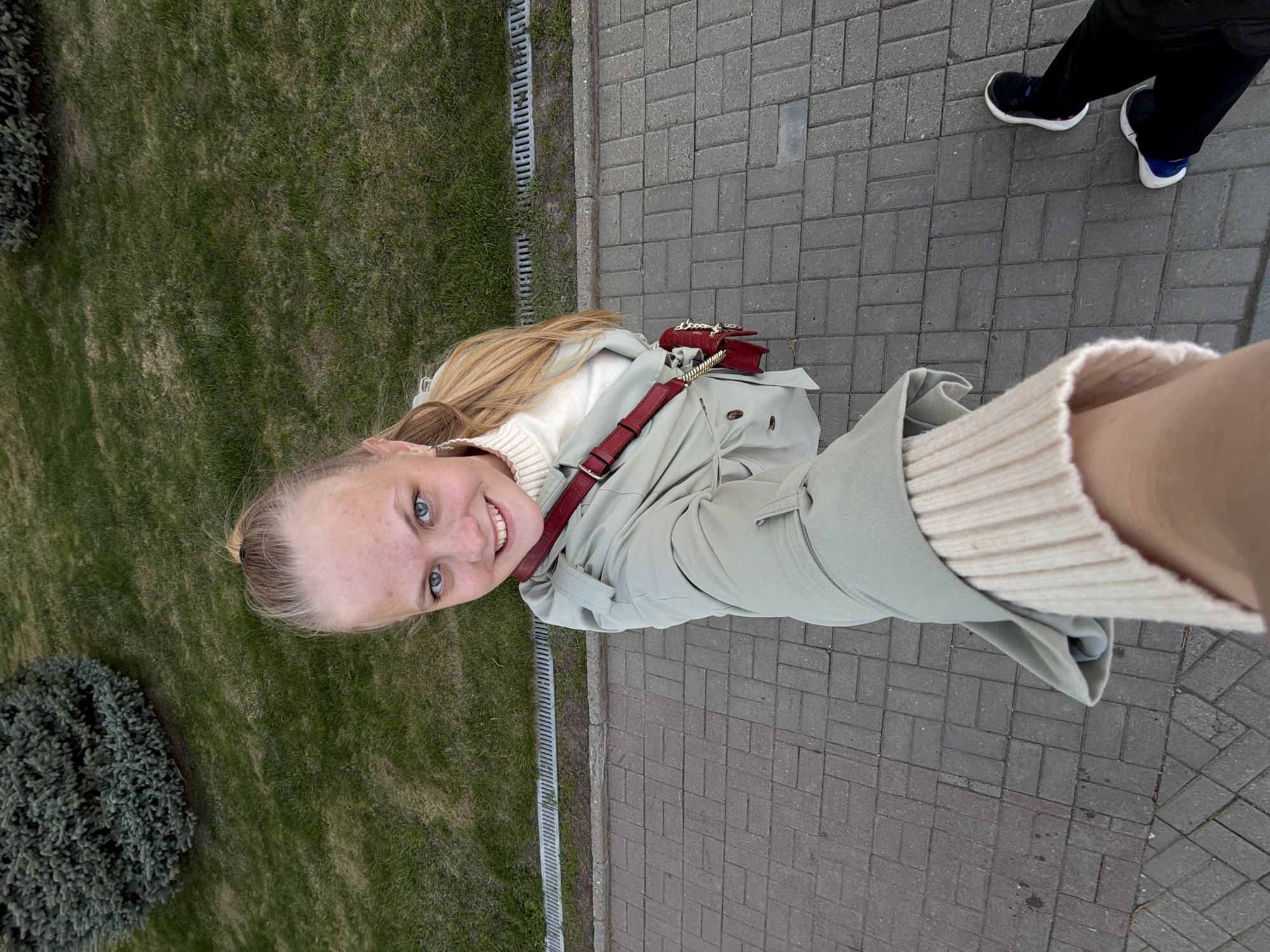 A woman is taking a selfie outdoors. She is standing on a brick-paved walkway next to a grassy area. The woman has blonde hair and blue eyes. She is wearing a white turtleneck sweater and a light green trench coat with a red purse strap. A second person is partially visible in the upper right corner of the image, wearing black pants and blue and white sneakers. The location may be in the city of Izhevsk, Russia.