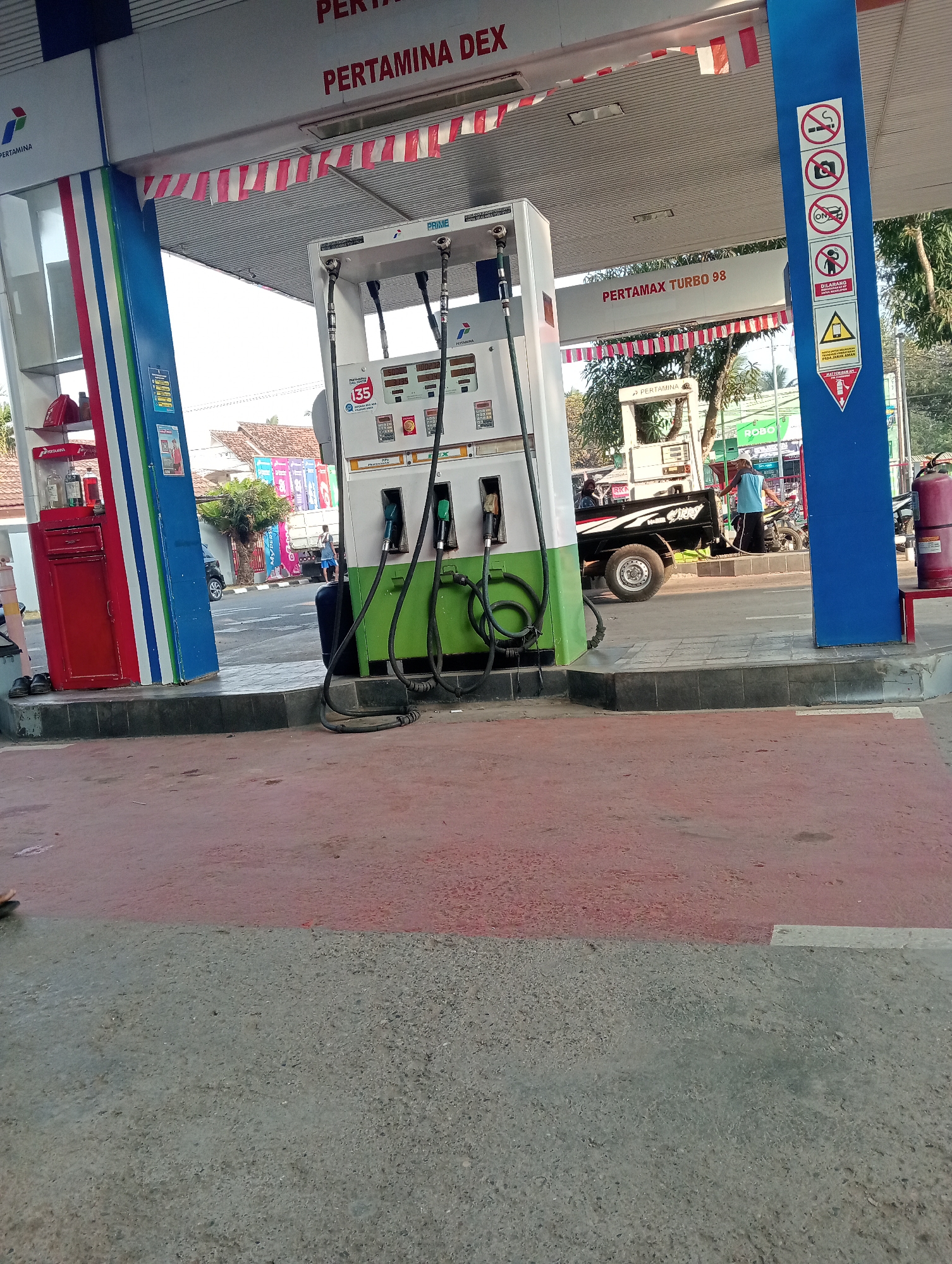 The image depicts a Pertamina gas station in Muara, Indonesia, on a bright, sunny day. The central focus is a white and green gas pump with hoses connected to it. Red and white Indonesian flags are strung across the awning of the gas station. On a blue pillar to the right of the gas pump, there are several warning signs, including one prohibiting smoking, one prohibiting photography, one indicating to turn off the engine, and another warning against opening flames and sparks. In the background, a black pickup truck is parked, and a person in a blue shirt is standing near it, possibly refueling a motorcycle. The ground in the foreground is paved with a red surface, transitioning to gray concrete. The overall environment suggests a typical day at a roadside gas station.