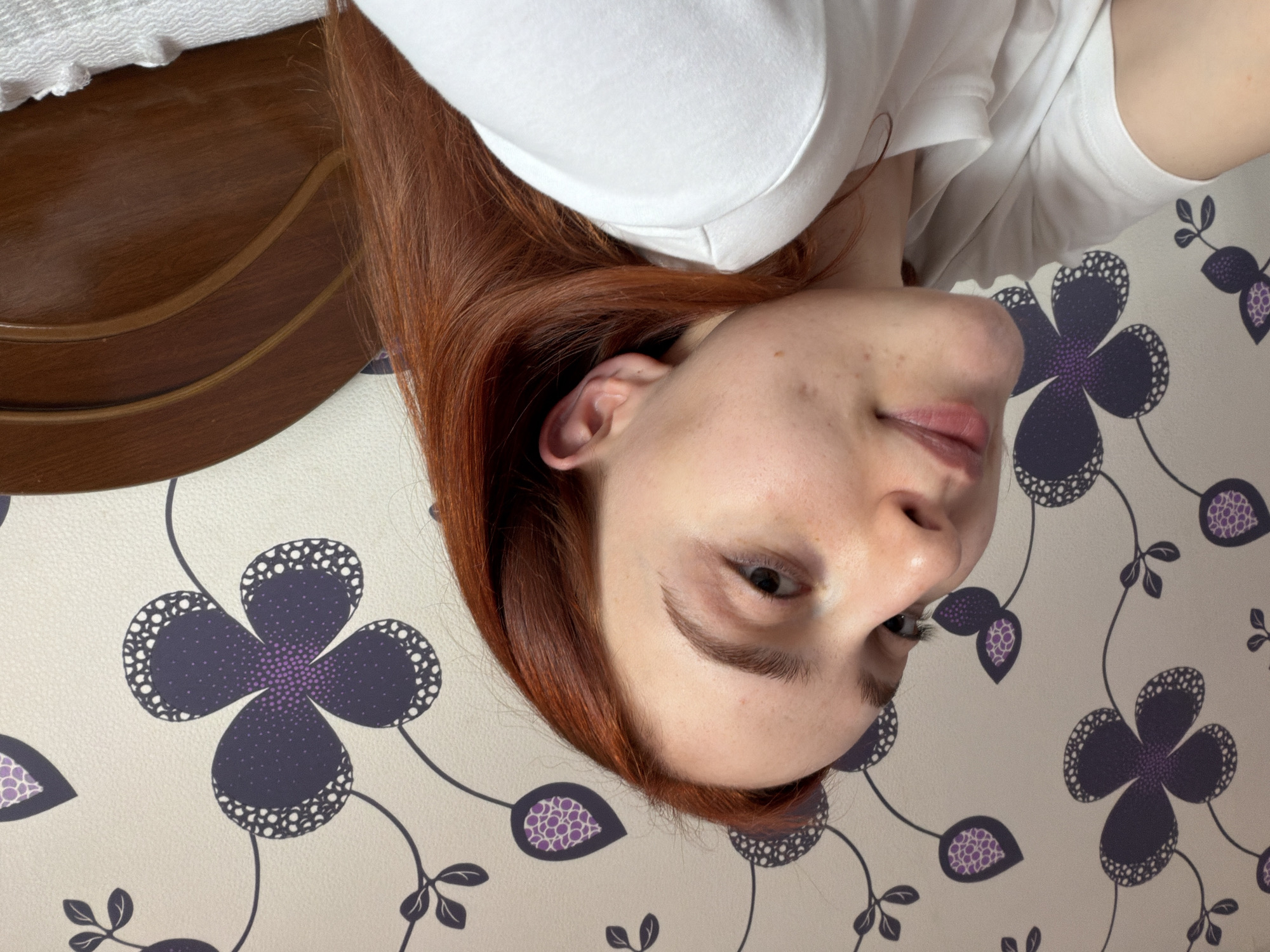 A person with red hair and a white t-shirt is lying on a bed with a wooden headboard. The person is on a bed with a floral-patterned wallpaper background. There is no evidence to suggest this photo was taken in Tbilisi, Georgia.