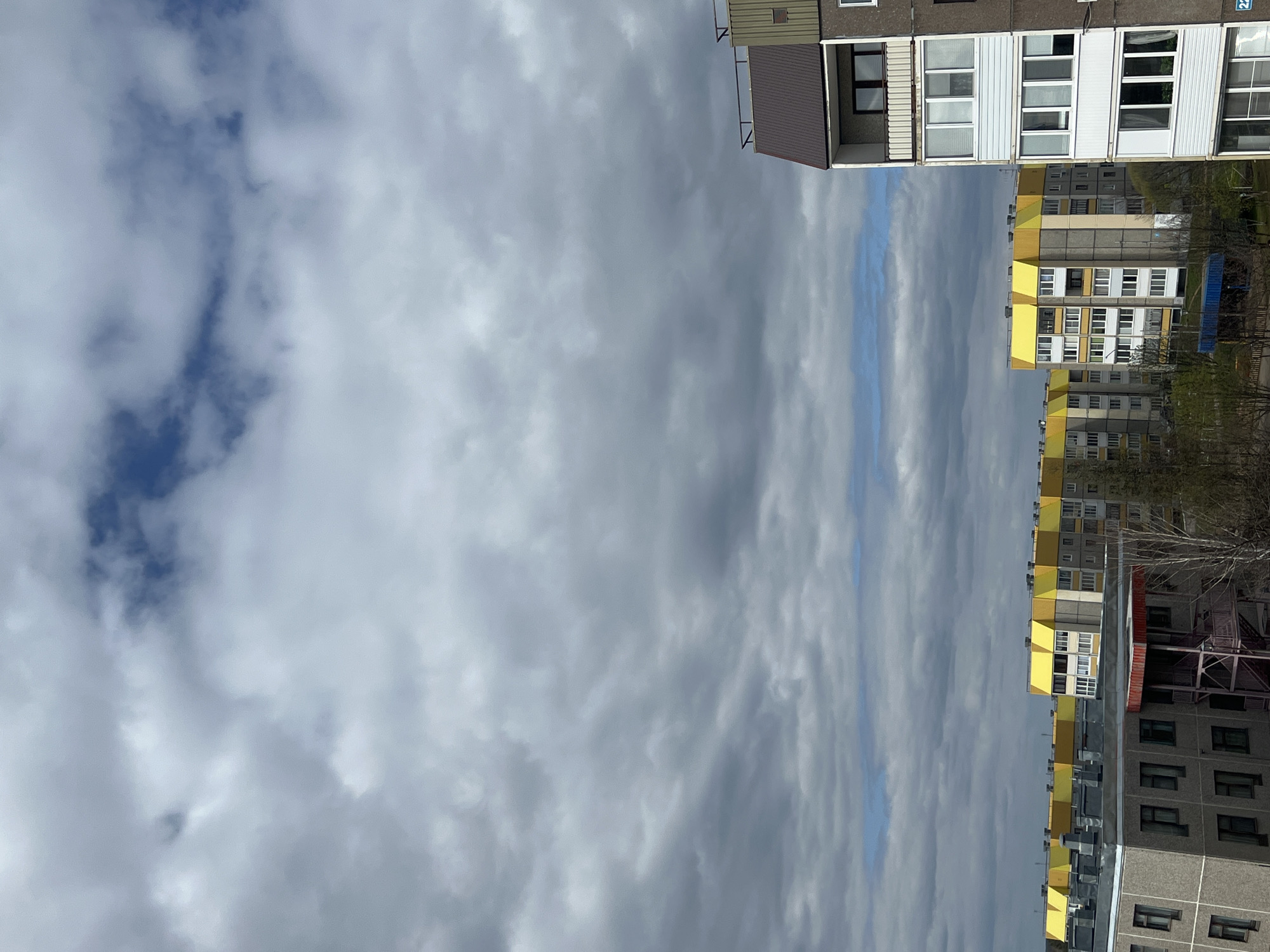The image depicts a cloudy sky over a city, possibly Марковский, Russia, as requested. The buildings are multi-story apartment complexes, with a mix of grey, white, and yellow exteriors. Some buildings have balconies. Trees can be seen in the background between the buildings. There are no people visible in the frame.