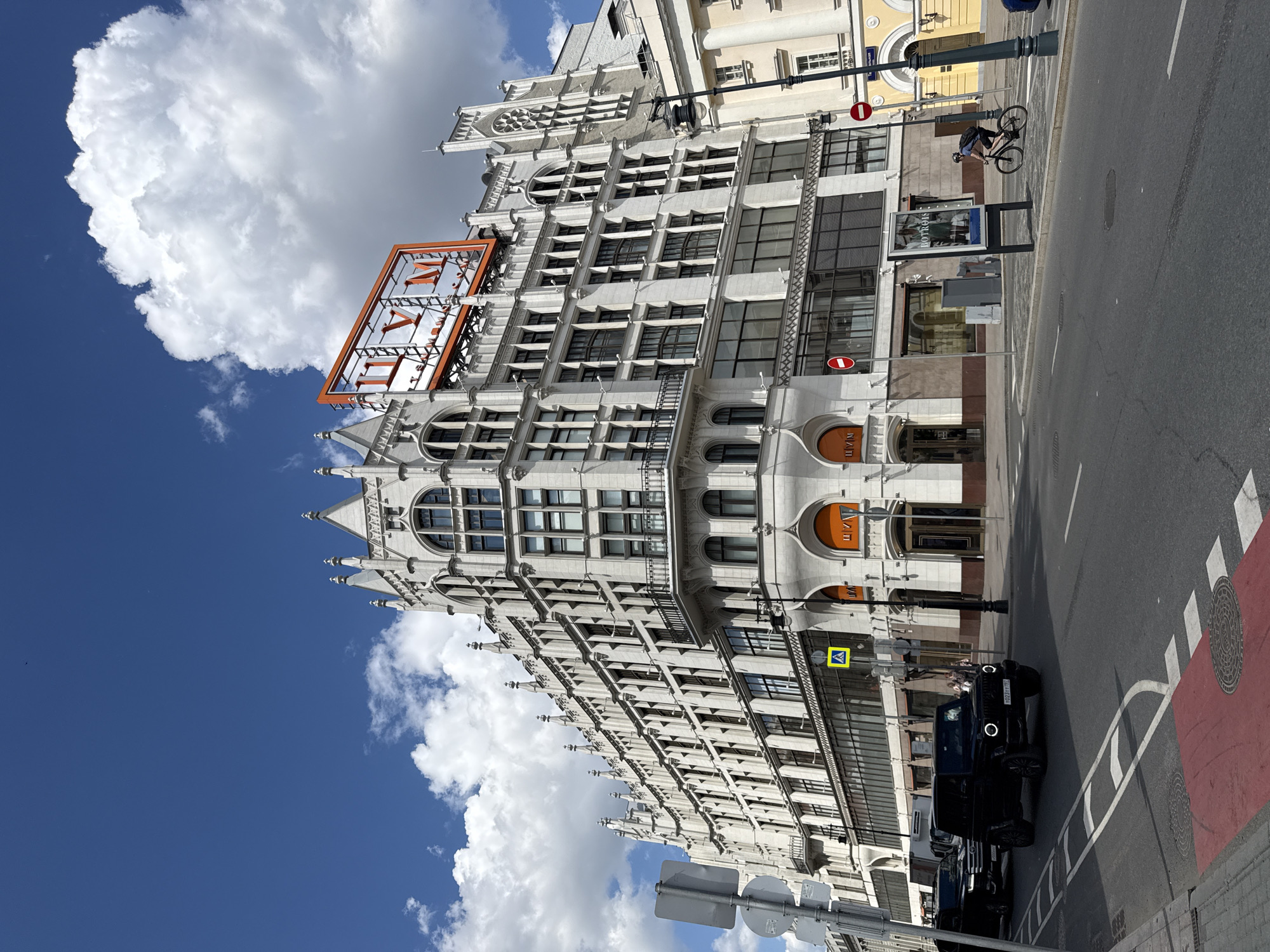 The image captures a vibrant daytime street scene in Moscow, Russia, under a bright blue sky dotted with large, fluffy white clouds. A majestic, ornate multi-story building dominates the left side of the frame. Constructed from light-colored stone or concrete, its architecture features numerous rectangular and arched windows, spires, and intricate Gothic or Neo-Gothic decorative elements along the roofline and façade.
Prominently displayed on the building's rooftop is a large, orange-framed sign spelling out "ЦУМ" (TSUM) in Cyrillic letters. Similar, smaller orange signs with "ЦУМ" are visible at ground-level entrances, identifying the building as a department store. An advertisement on a street-side banner near the pavement shows the text "MONCLOU," likely referring to the luxury brand Moncler.
The street itself, occupying the right side of the image, is made of asphalt and features various road markings, including distinct red and white striped sections, suggesting a bus lane or pedestrian crossing, and white lane lines. A black Mercedes-Benz G-Wagen SUV with a visible Russian license plate "Y023PH 799 RUS" is on the road, with other vehicles partially visible further down the street.
A street lamp post, various traffic signs, including a yellow diamond-shaped sign (pedestrian crossing ahead) and red "No Entry" signs, are also present. One person, a cyclist wearing a helmet and backpack, is seen riding a bicycle on the sidewalk adjacent to the road. The clear sky and bright lighting indicate a sunny day.