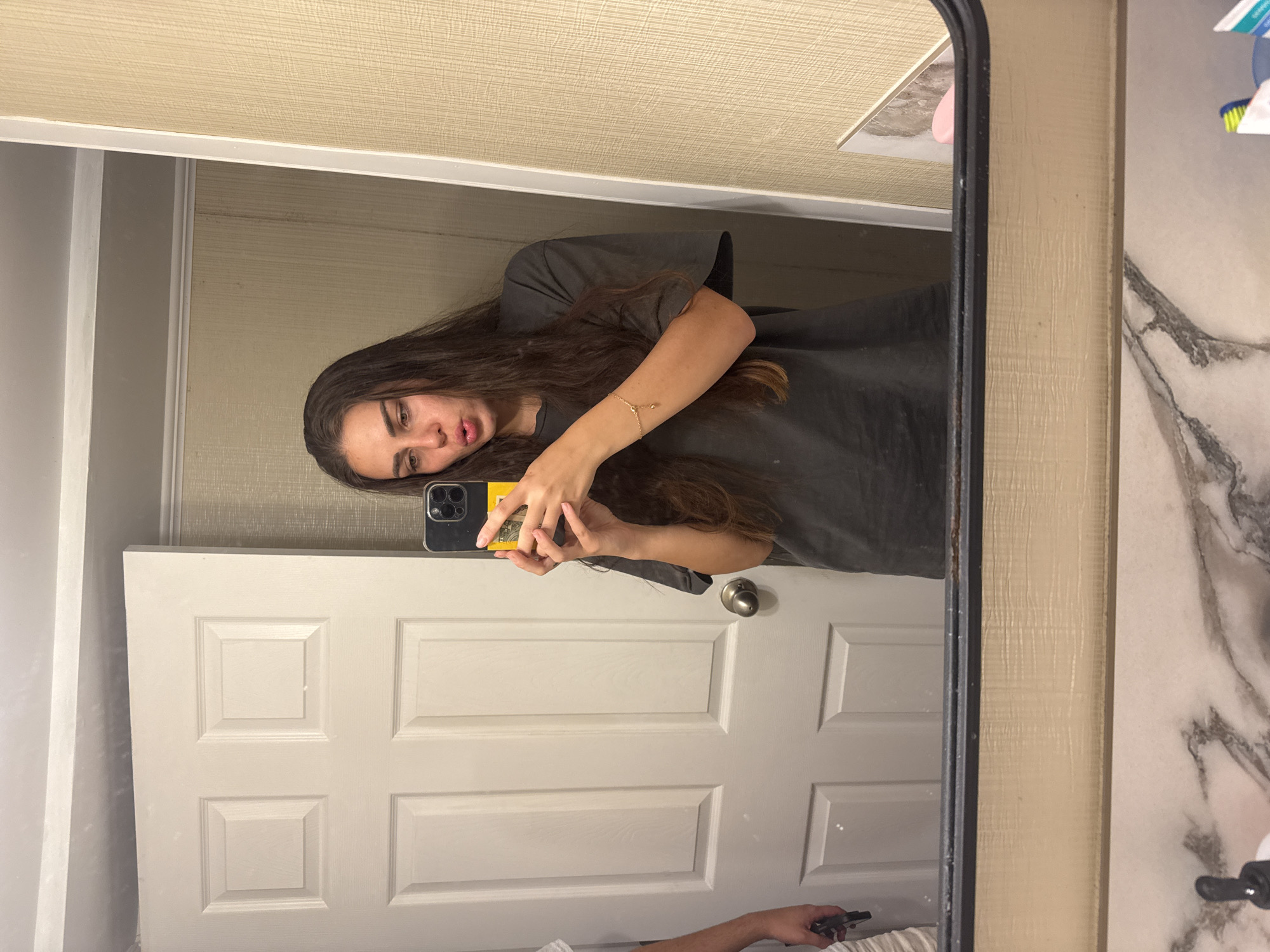 A young woman with long, dark hair and an olive complexion is taking a mirror selfie in a bathroom. She is wearing a dark gray t-shirt and a delicate gold bracelet on her left wrist. Her lips are pursed, and she is holding a smartphone with a yellow case in her right hand to capture the reflection. The background shows a white paneled door and a textured beige wall. The bottom of the image displays a portion of a marble-patterned countertop with various toiletries, including a pump bottle, a pink object, and a tube of toothpaste. The overall scene suggests a casual moment in a domestic setting. The specific location of Ocean City, United States of America is not discernible from the provided image.