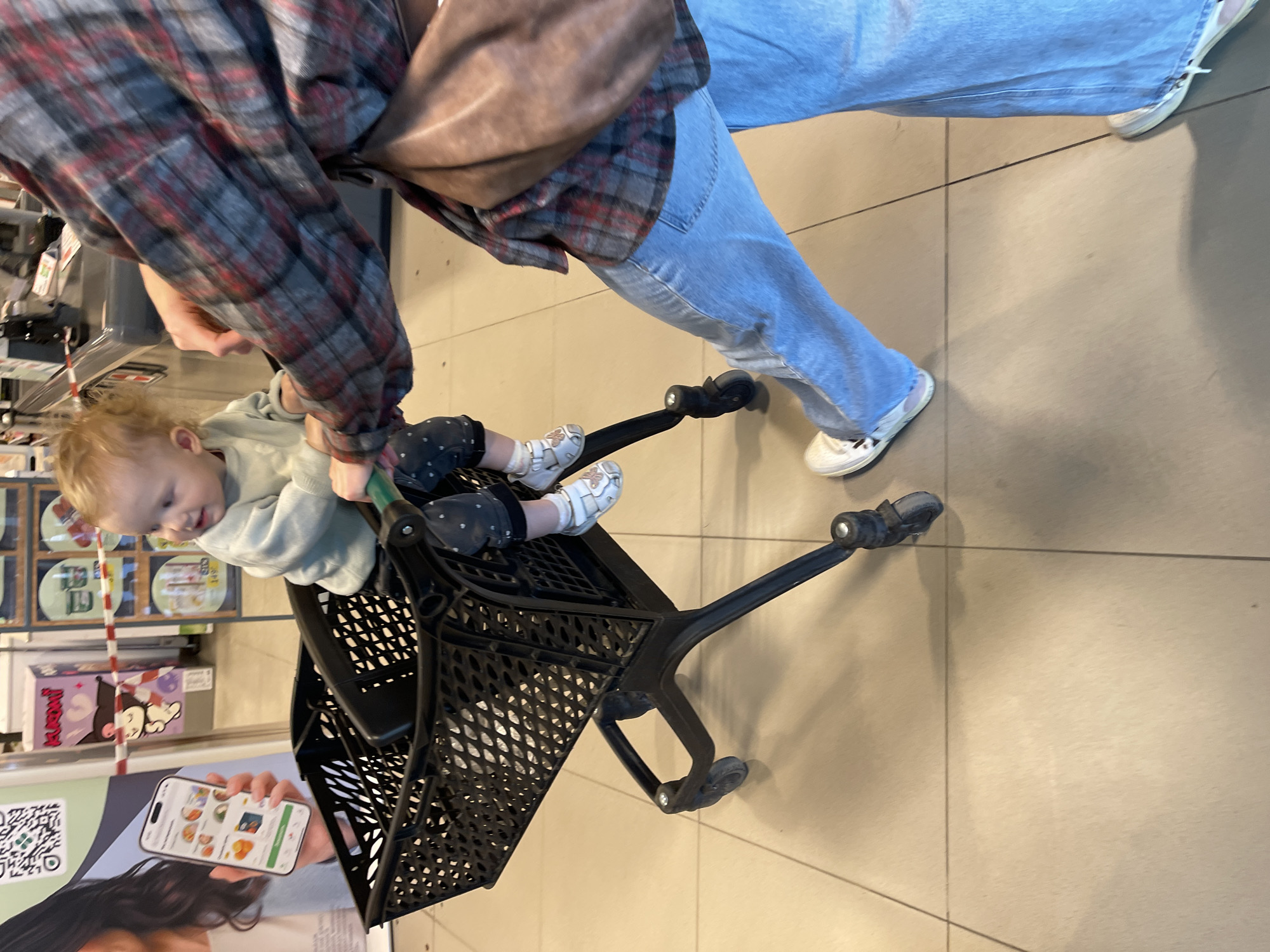 An indoor scene, rotated 90 degrees counter-clockwise, shows an adult pushing a shopping cart containing a toddler, likely within a supermarket in Moscow, Russia. The adult, visible from the waist down and arm, wears a red and grey plaid shirt, blue jeans, and white sneakers, with a brown shoulder bag. Their hand holds the handle of a black, plastic shopping cart.
A young child, with light, possibly blonde, curly hair, sits in the cart's child seat. The toddler wears a light green long-sleeved top, dark patterned pants, and white and beige sandals, looking towards the left with a content, slightly smiling expression while gripping the cart handle.
The setting features light-colored tiled flooring and store shelves lined with products. To the left of the child, a large advertising poster displays a hand holding a smartphone with a grocery or food delivery app interface, featuring food images and a clear QR code. Text on product packaging on the shelves includes "KitKat" on a purple box and "KiKoMi" on a white box. The image captures a moment of a parent and child shopping.