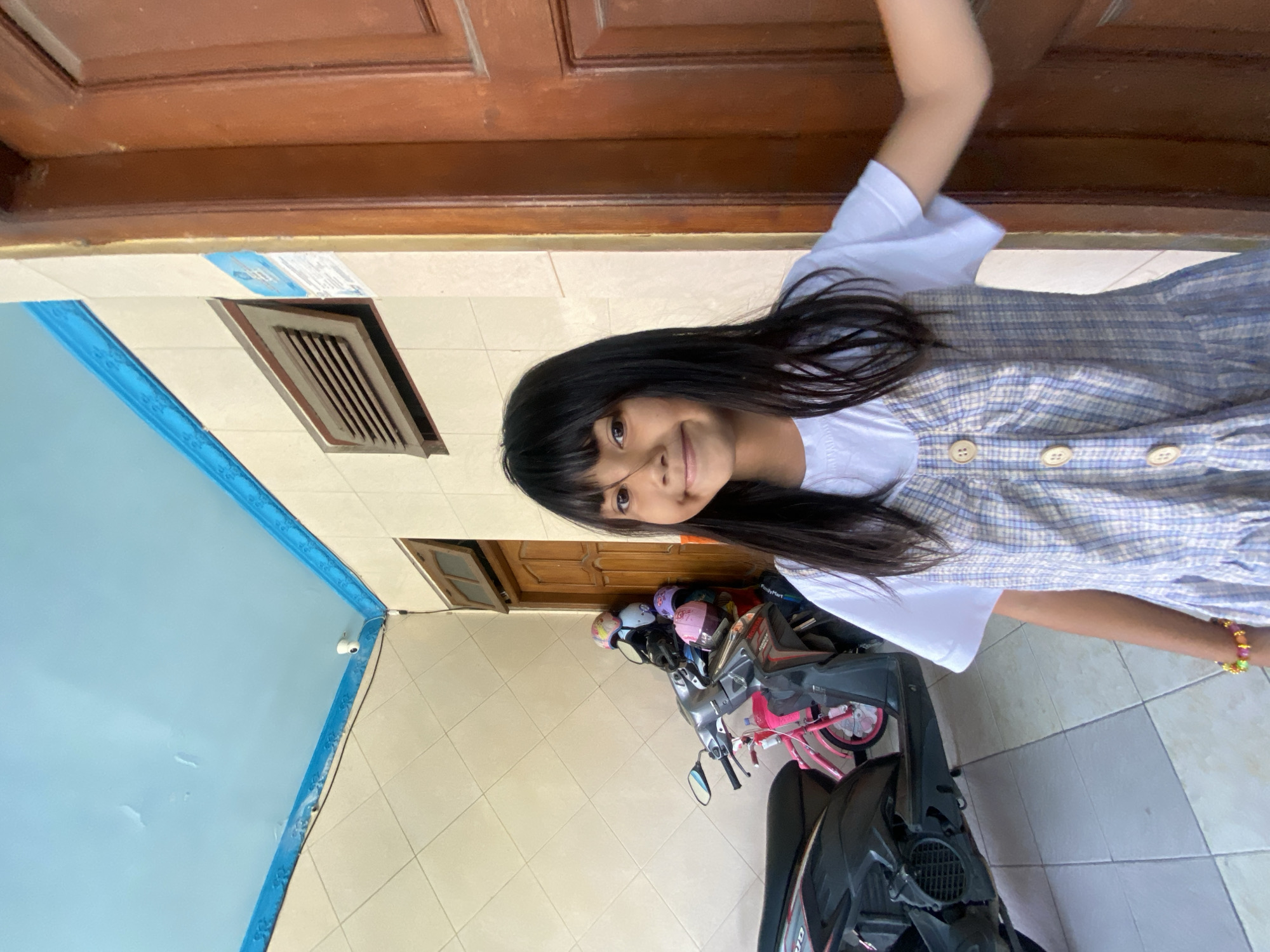 A young girl with long, dark hair and bangs is smiling at the camera. She is wearing a white t-shirt under a blue and white plaid dress. She has a bracelet on her left wrist. The girl is standing in front of a wooden door on the right side of the image. To her left, there are two parked motorcycles and a wall with a vent. The ceiling is light blue with a blue border. The floor is tiled. The environment appears to be an outdoor porch or entryway. This scene is located in Gelam, Indonesia.