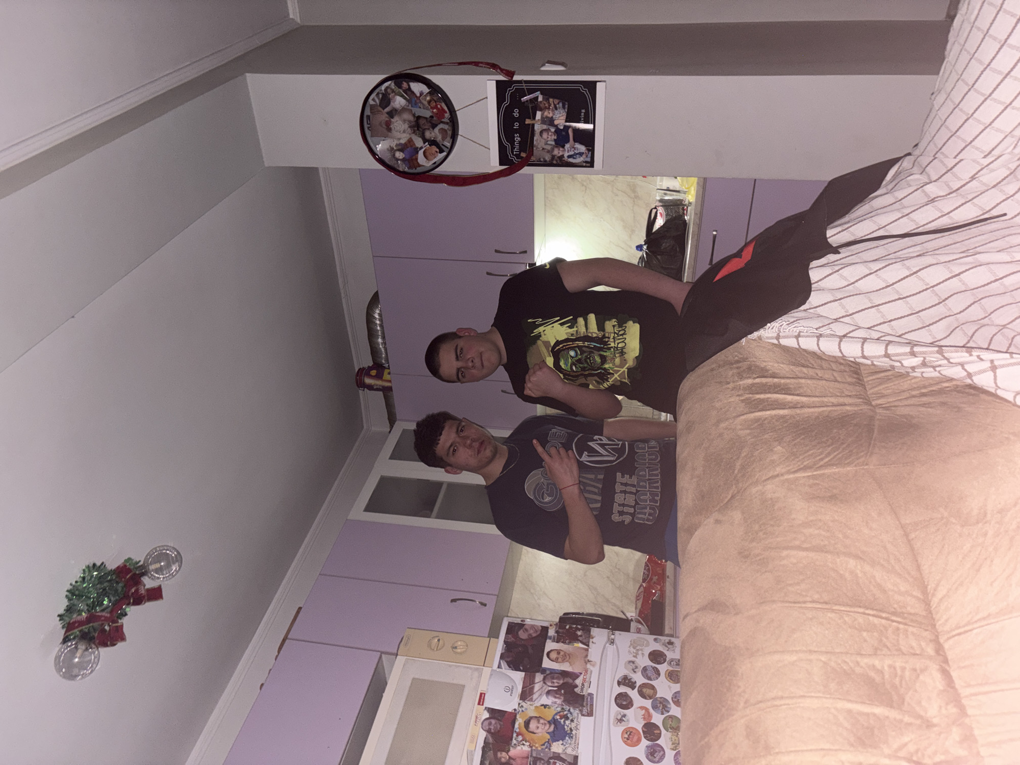 Two young men are indoors, possibly in a residence. One is wearing a dark t-shirt with a green graphic, the other a dark t-shirt with a lighter graphic. The setting includes kitchen cabinets, a refrigerator adorned with photos and magnets, and furniture, possibly a couch. The décor includes a wreath on the ceiling and wall hangings above the cabinets. The city and specific location are unknown.