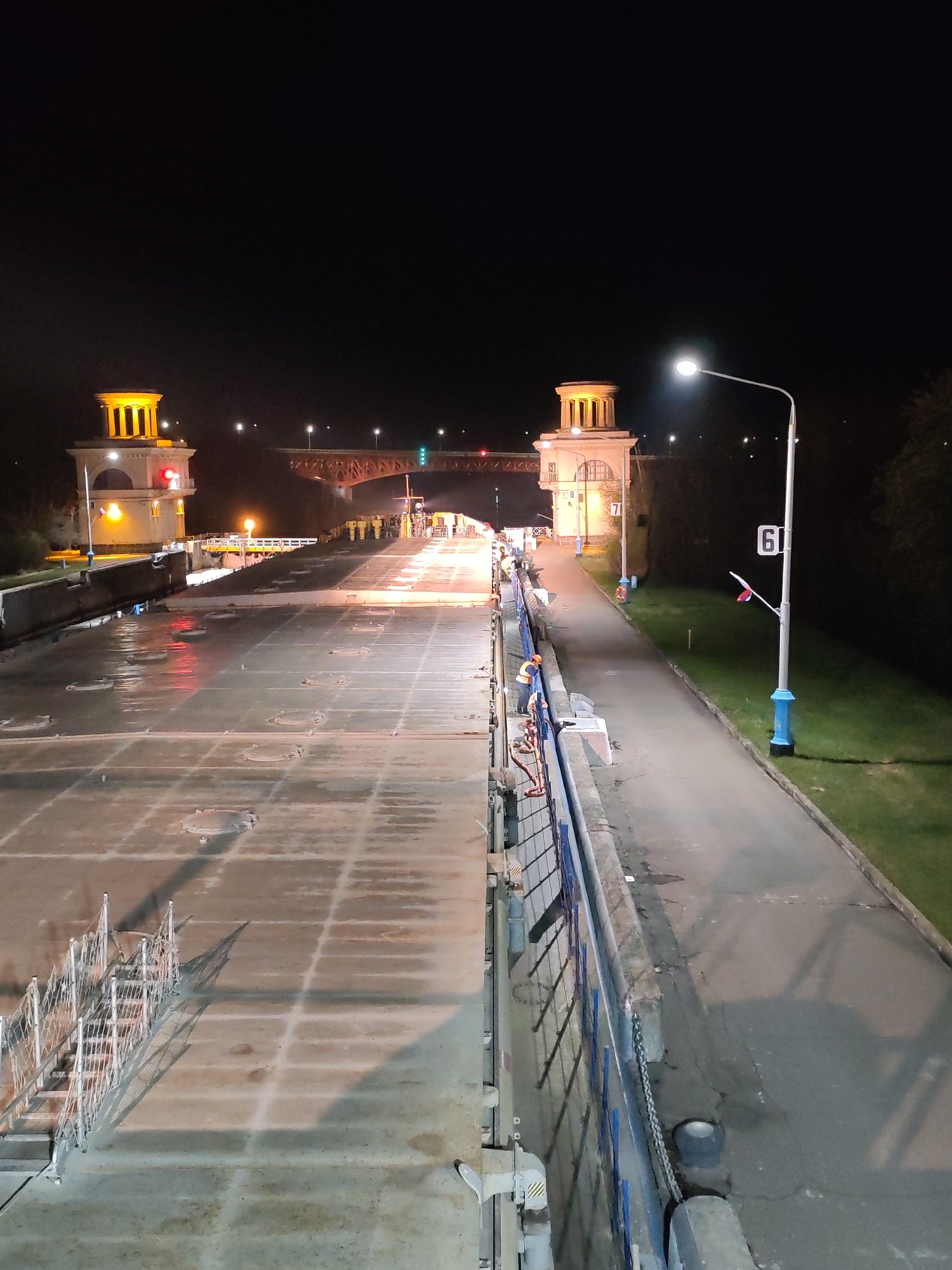 The image depicts a night scene at the lock connecting the Volga and Don rivers at Базарово, Russia. The lock appears to be closed and empty of water. On either side of the lock chamber are control towers lit with yellow lights. In the distance, a bridge with red and green lights spans the waterway. A street lamp illuminates a paved path alongside the lock. A sign labeled "61" is next to the street lamp. Along the lock's edge, a blue metal railing can be seen. A white metal staircase leads down to the floor of the lock. The overall impression is of an industrial or engineering site at rest during the night.
