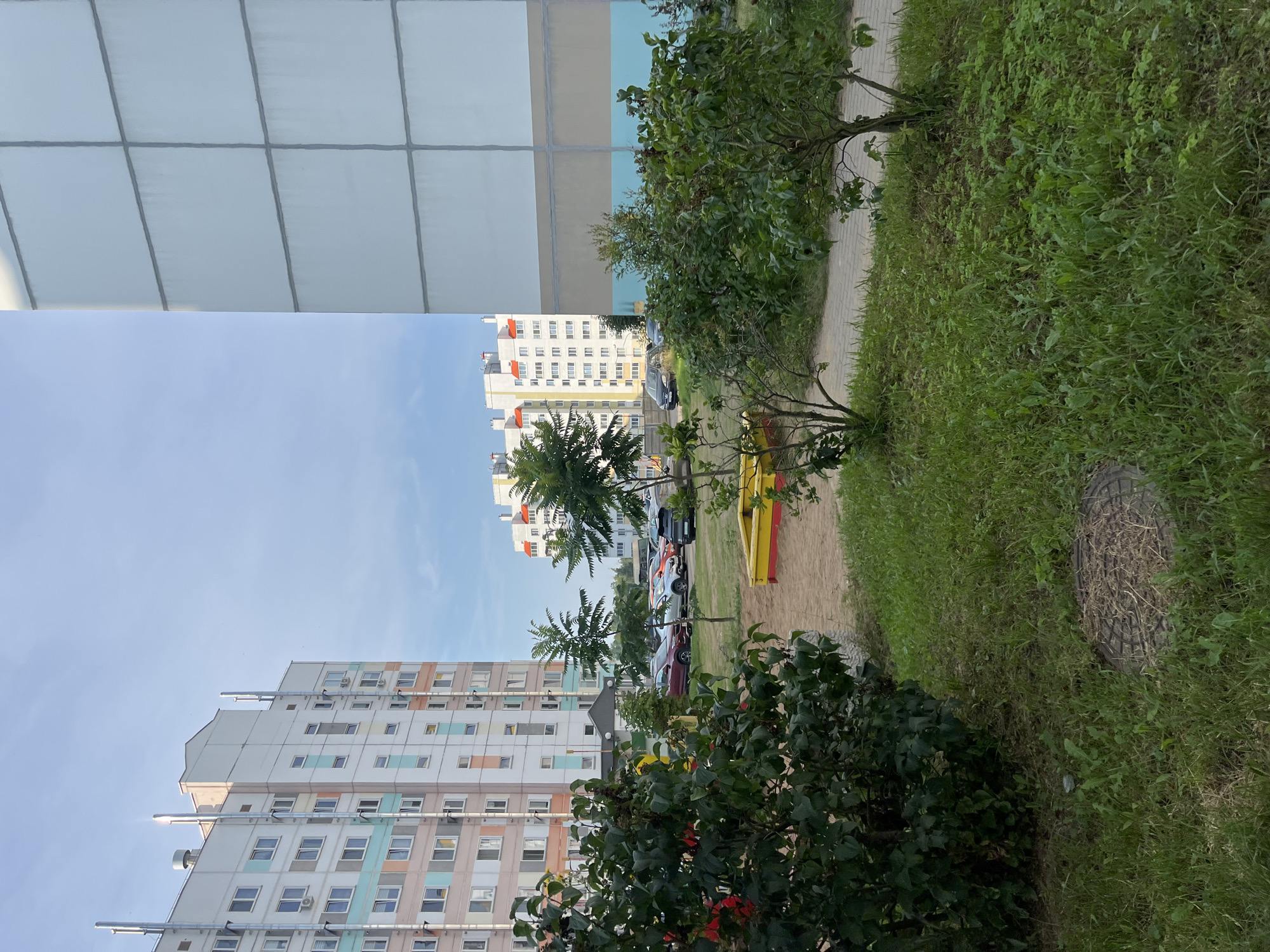 This is a daytime, eye-level exterior shot of a residential area in the Senitsky Sel'skiy Sovet, Belarus. The scene is dominated by multi-story apartment buildings. On the left, a light-colored building with pastel accents (pink, blue, and white) rises against a clear blue sky. Several prominent metal pipes run vertically along its facade, suggesting ventilation or exhaust systems. In the foreground of this building's side, dense green bushes with red flowers are visible.
To the right, another apartment building is partially visible. Its facade is a large, uniform light gray, divided into square panels by dark gray lines. A horizontal band of light brown is visible below a band of light blue, about two-thirds of the way up the building.
Between these buildings, a playground area is evident. A yellow and red sandbox is situated on a sand-covered patch. Several trees are scattered throughout the scene, some with sparse foliage. Cars are parked in a lot behind the sandbox and playground. The ground is covered with a mix of grass and a paved walkway. A manhole cover is visible in the grassy area on the left side of the frame. The overall impression is of a typical, somewhat bland Soviet-era residential block.