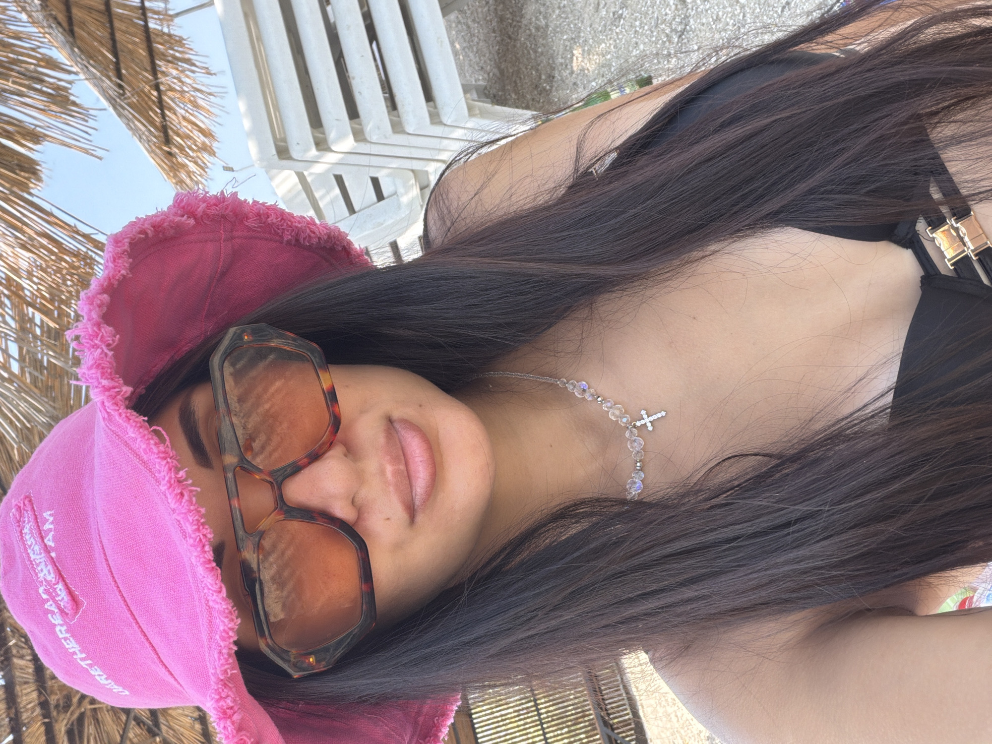 In an outdoor, beachfront setting, a person is taking a selfie. The subject is wearing a bright pink bucket hat with frayed edges, large, tortoiseshell-patterned sunglasses with amber lenses, and a black bikini top with gold hardware. A delicate necklace with clear beads and a small silver cross pendant adorns their neck. Their long, dark hair is visible, framing their face and falling over their shoulders. In the background, beach chairs and umbrellas are visible, suggesting a sunny day at the beach. The location is identified as Pavlo-Ochakovo, Russia.