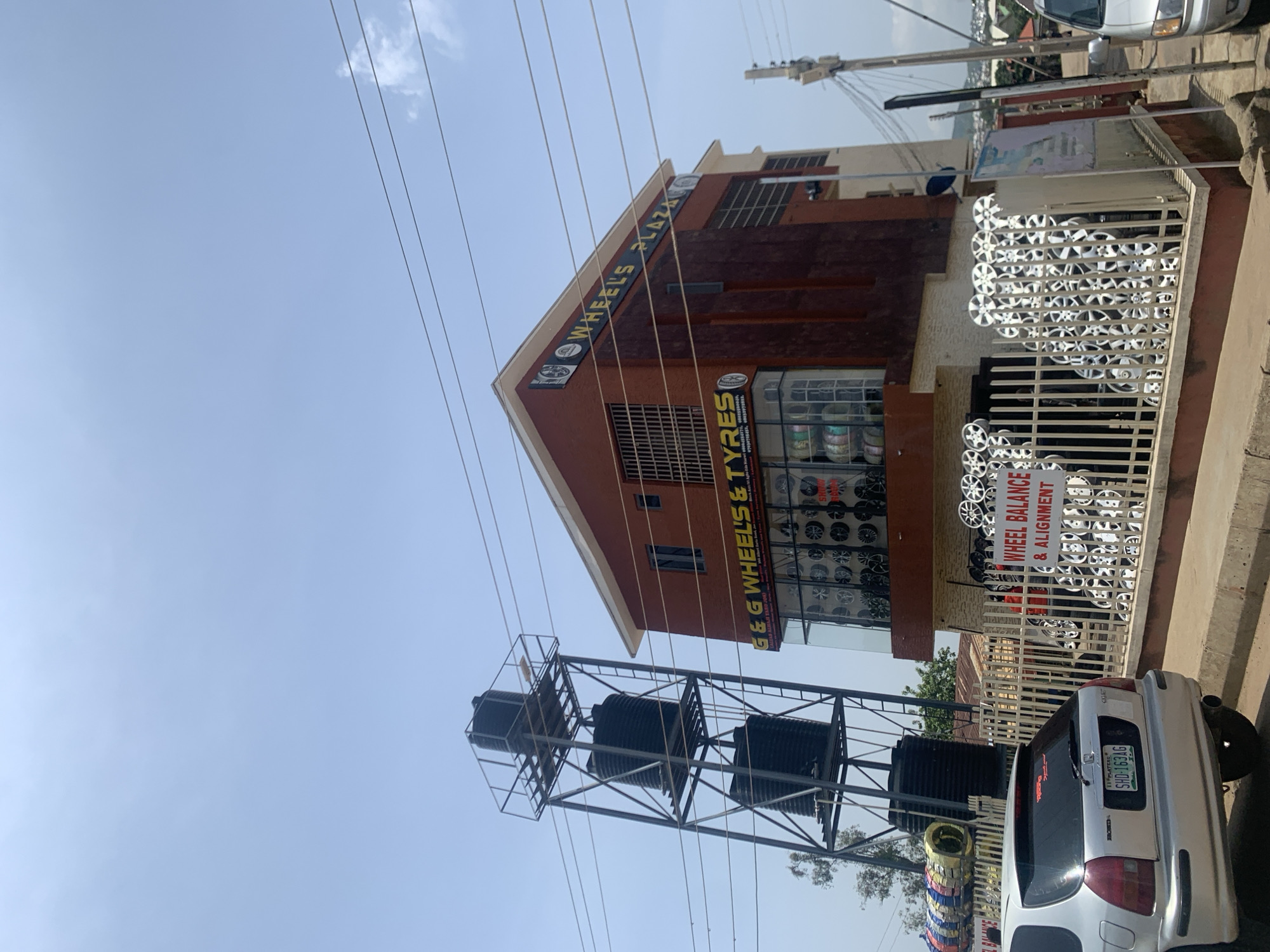 In the city of Jos, Nigeria, a building with a red brick facade stands next to a tall, industrial metal structure supporting three black water tanks. The building houses "G & G WHEELS & TYRES" and "WHEELS PLAZA," with a display of car wheels visible through a glass storefront. In front of the building, a metal fence encloses a display of numerous car wheels, some arranged in a decorative pattern. A silver Honda Civic car is parked on the left side of the scene, facing away from the camera, with its license plate reading "SHD-163AG." The sky above is a pale blue with a few wispy clouds, and electrical wires are strung across the frame. The environment appears to be a commercial street.