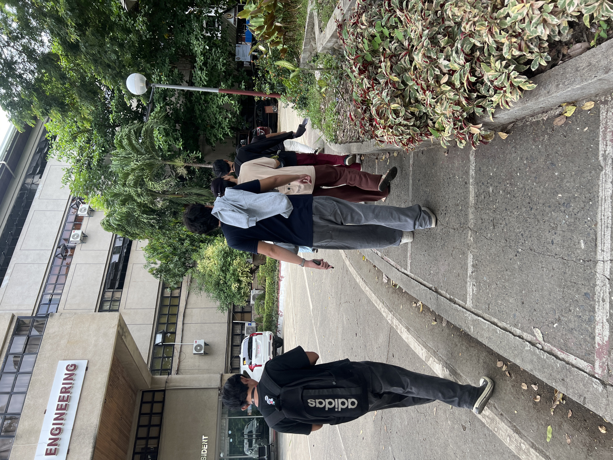 A medium-angle outdoor shot depicts a group of people standing in a line next to a building on a partially overcast day.
A group of five people are positioned in a row, on the sidewalk, with their backs to the camera. The person closest to the camera is wearing a black shirt, dark pants, and a black Adidas backpack. Progressing along the line, others are wearing a dark shirt and light gray pants, a beige shirt and maroon pants, a cream-colored shirt and maroon pants, and a black shirt and pants. They are standing along a gray concrete sidewalk next to a parking area.
To the left of the line, a building with a sign that reads "ENGINEERING" is visible. Air conditioning units are mounted on the building's exterior. A white van is parked nearby. Green trees and bushes are visible behind the people and along the sidewalk, as well as a street light.
Based on the building visible with "ENGINEERING" sign, the location is likely in Davao City, Philippines.