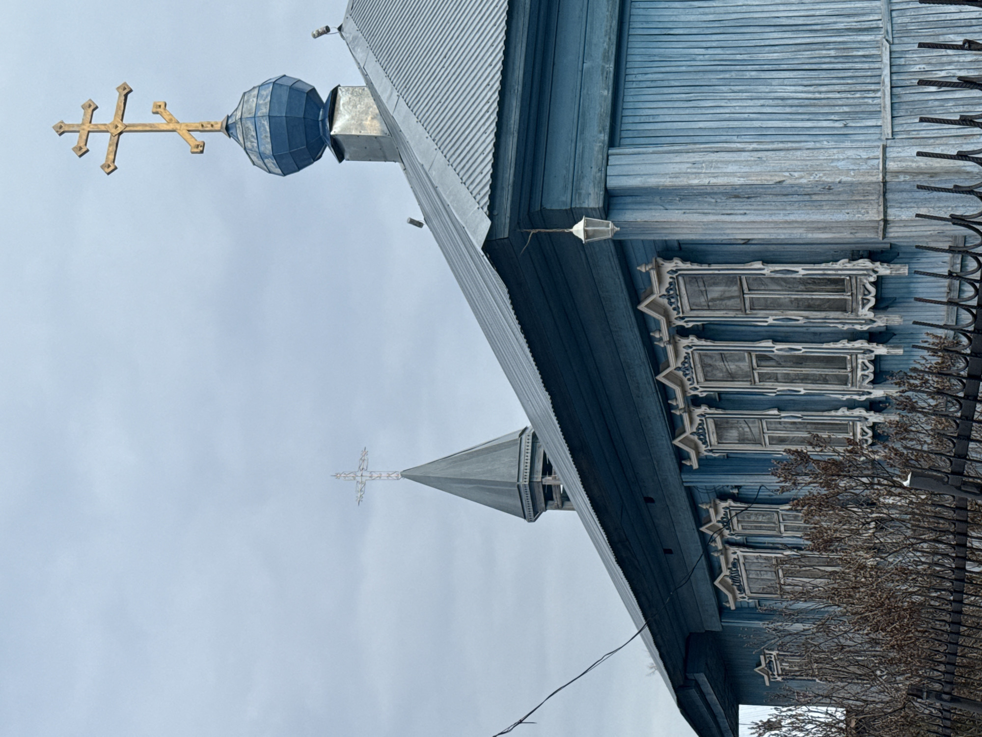 A tilted low-angle shot depicts a blue wooden building, possibly a church, against a cloudy sky. The architectural features include a dome with a golden cross, ornate window frames, and a pointed steeple with another cross. A black metal fence and bare tree branches partially obscure the building's side. The location is Фоки, Russia.