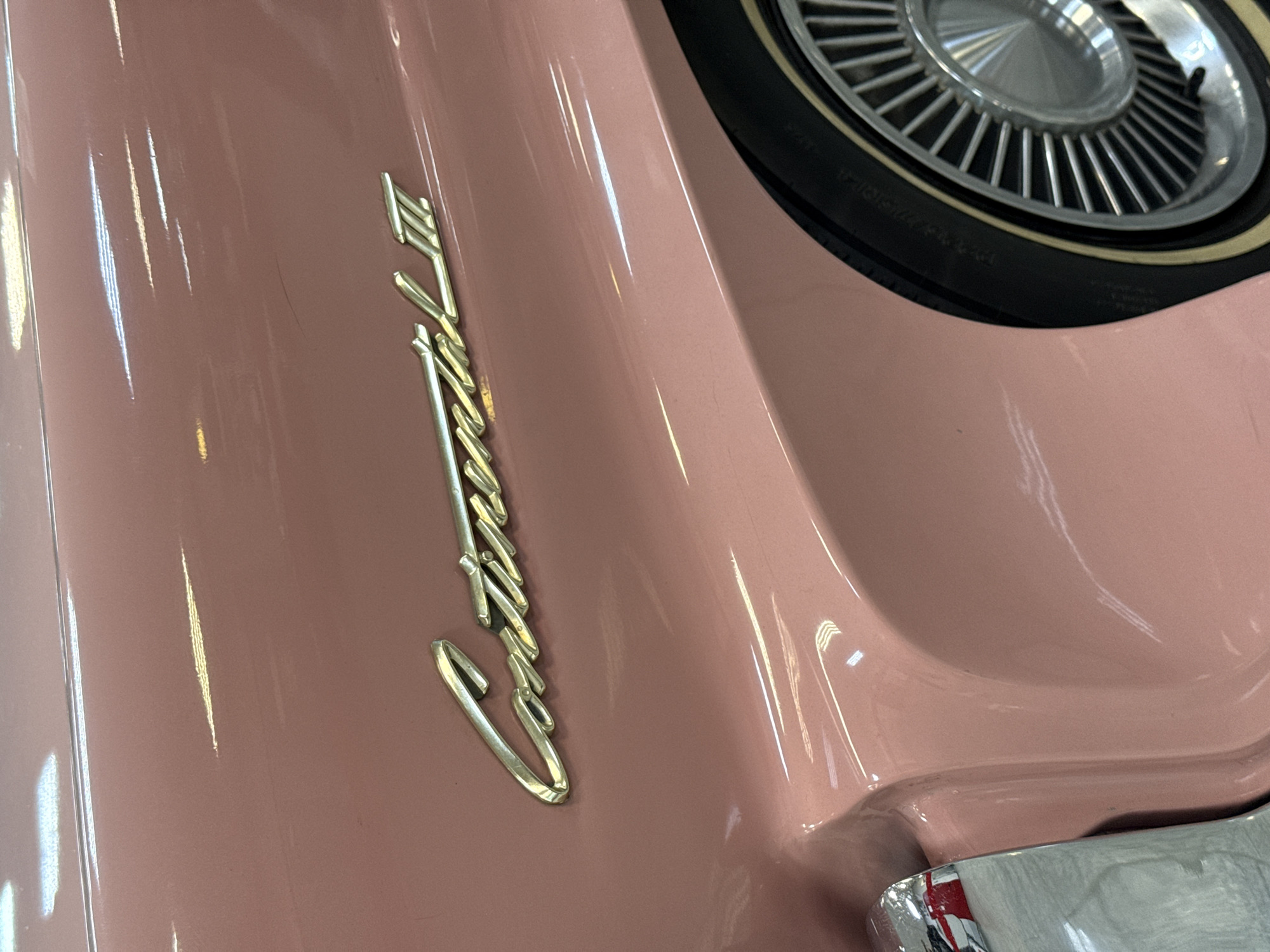 This close-up image captures a portion of a vintage car, showcasing its side panel and rear wheel. The car is painted in a distinctive, glossy light pink or coral hue, which is highly reflective, showing elongated streaks of indoor lighting.
Prominently featured on the side of the car is a gold-colored, script-style emblem that clearly reads "Continental Mark II," indicating the model of the classic vehicle. Below this text, the body curves gently, and towards the bottom right, a small part of a chrome bumper is visible, reflecting a distorted pattern of red, white, and blue, possibly from a flag.
The upper right section of the image shows a white-wall tire and a multi-spoked chrome hubcap, typical of classic automobiles. The tire sidewall has some indistinct, unreadable text. The overall scene suggests the car is meticulously maintained and is likely displayed indoors, perhaps in a showroom, museum, or private collection in Sochi, Russia. No human subjects or specific activities are depicted; it is a static shot highlighting the car's aesthetics and details.