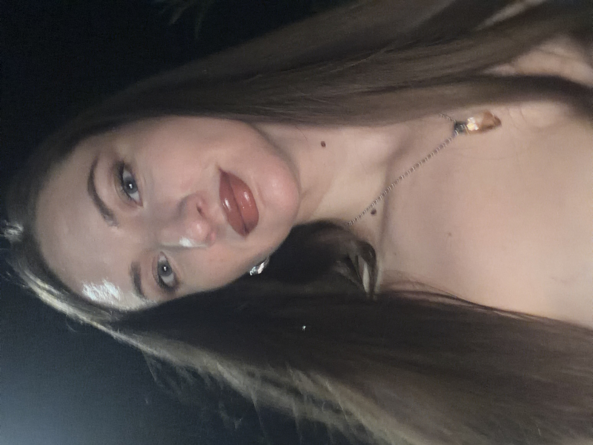 A woman with long, dark brown hair is captured in a close-up, low-angle selfie outdoors at night. She is wearing lip gloss and a silver necklace with a crystal pendant. Her head is tilted slightly, and she is looking towards the camera. The background is dark, suggesting an evening setting, possibly in a garden or public area. The image quality is slightly grainy. The location is identified as Sharm El Sheikh, Egypt.