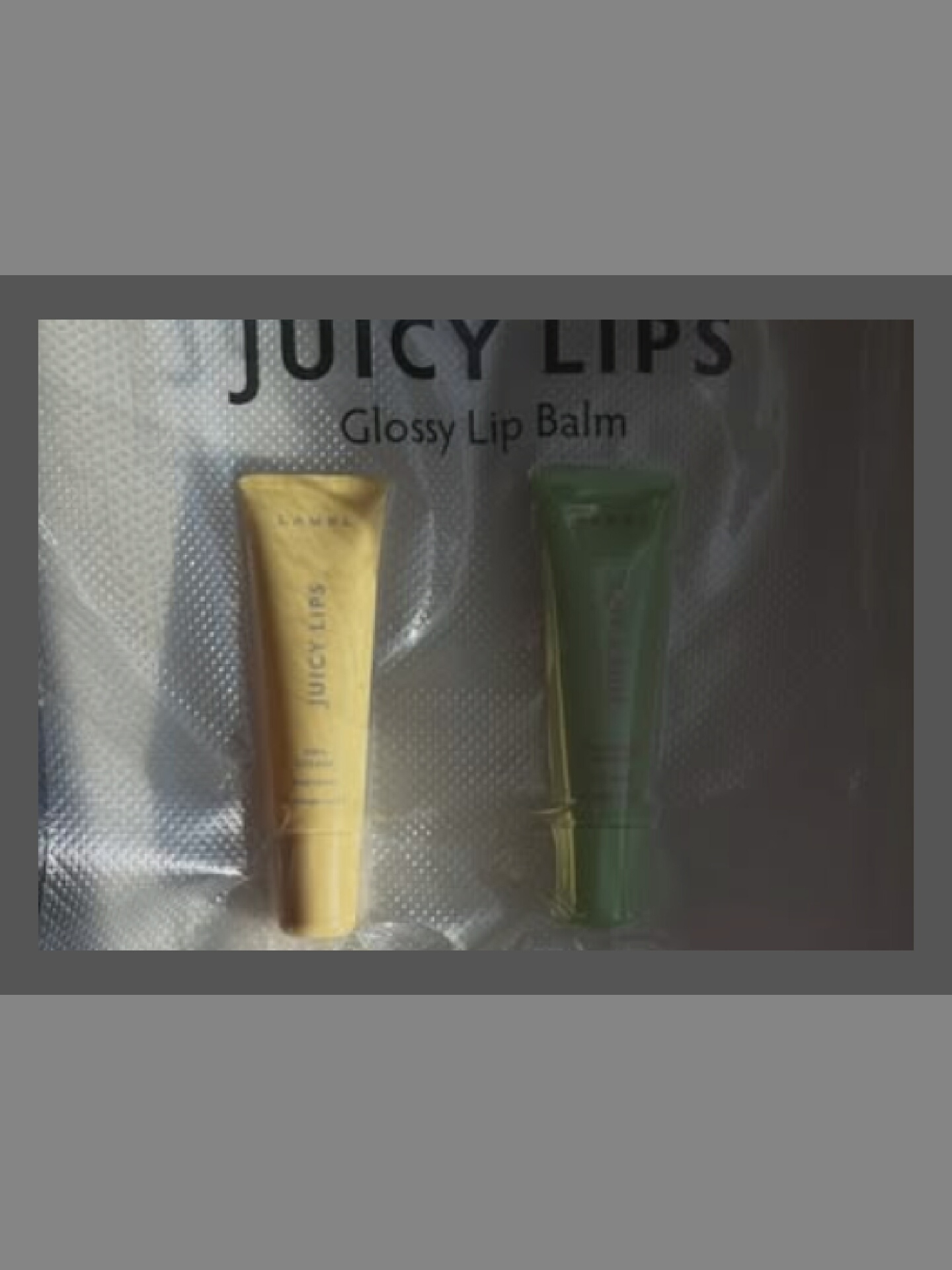 Two tubes of lip balm are displayed in front of a reflective, textured background. The tube on the left is a pale yellow and features the text "JUICY LIPS" vertically on its side, along with a logo that appears to be "L'AMEL". The tube on the right is green and also appears to have text, but it is not clearly legible. Above the lip balm tubes, the words "JUICY LIPS" are printed in large, black, capitalized letters, with "Glossy Lip Balm" printed in smaller font below. The overall presentation suggests a product advertisement or display.