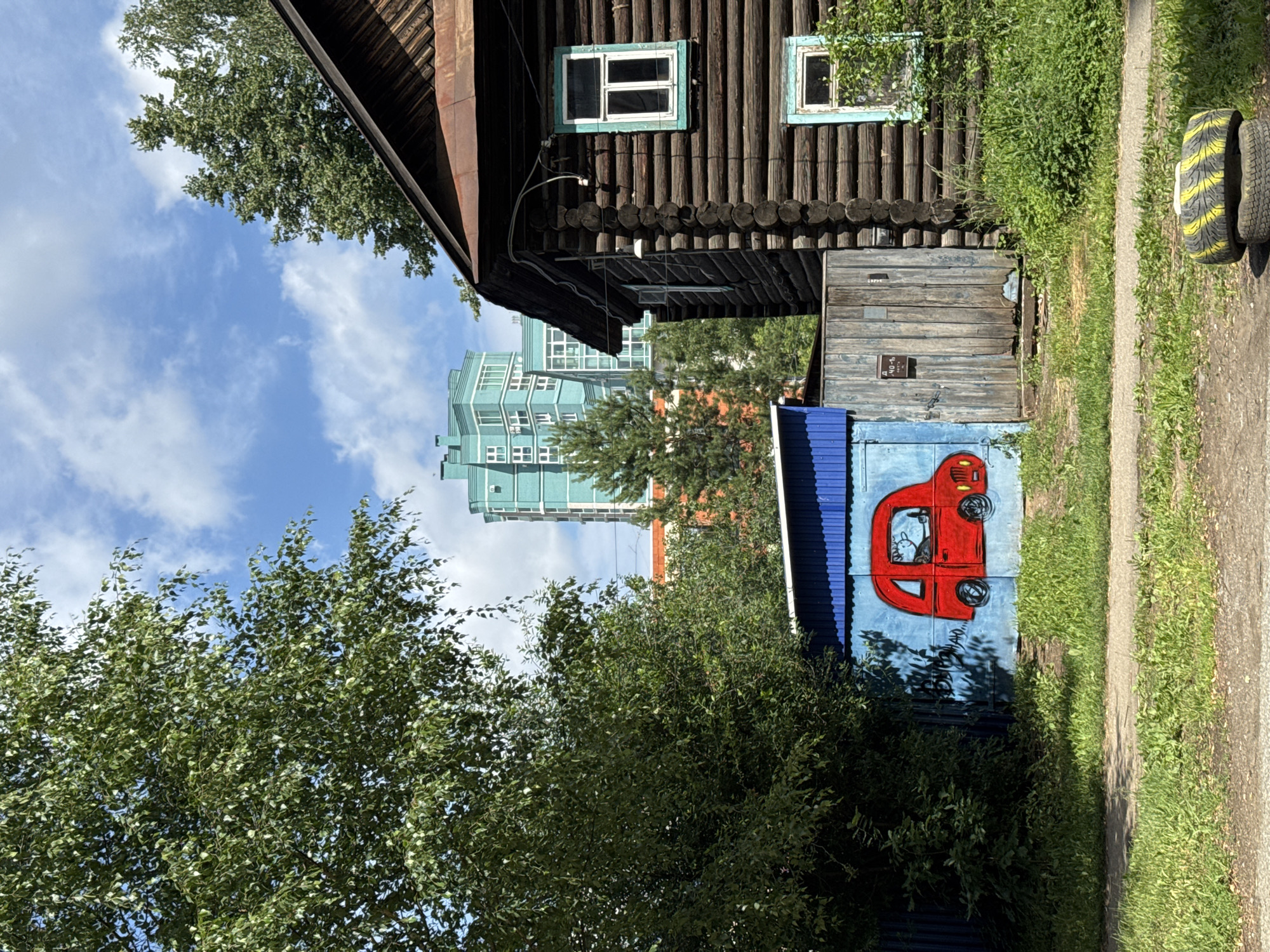 The image depicts a scene in the city of Izhevsk, Russia. In the foreground, on the right, is a weathered wooden house with visible logs and a rusty metal roof. It has two windows with light blue trim. To the left of the house is a blue corrugated metal structure with a faded mural of a red car. The car mural has a simple drawing of an animal figure inside, possibly a dog. A wooden gate is situated between the house and the blue structure, showing signs of wear. Overgrown green grass and vegetation are present throughout the scene. In the background, a modern, tall, teal-colored building with many windows stands out against a blue sky with scattered white clouds. Trees are visible throughout the image, with a large leafy tree dominating the left side. The overall impression is a juxtaposition of older, rustic structures with newer urban development.