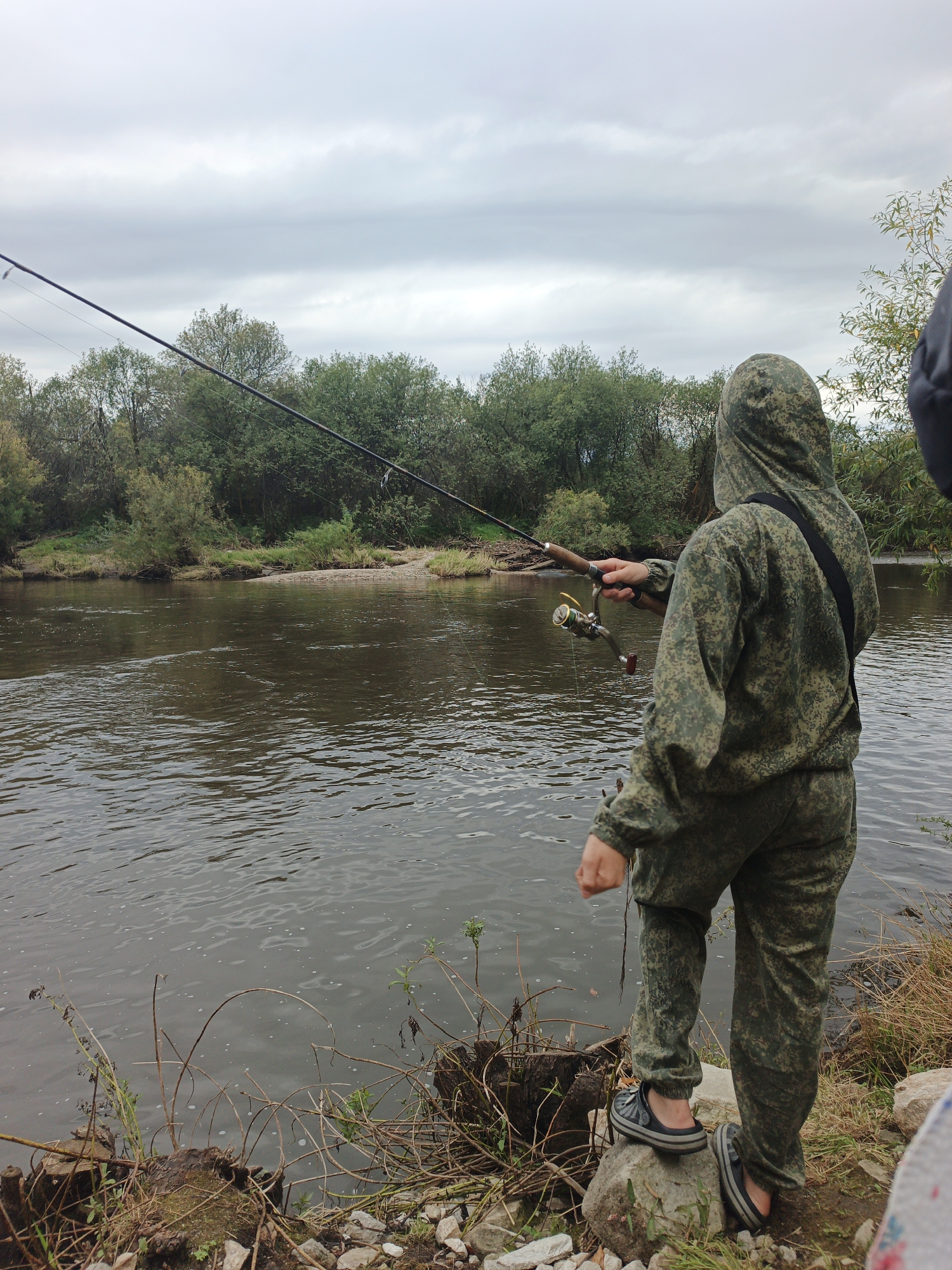 A person in camouflage fishing attire stands by the muddy bank of a river, fishing with two rods. The person is facing away from the camera, looking out at the water. The river is wide and calm, with trees and vegetation lining the opposite bank. The sky is overcast. The scene appears to be in Medvedyovka, Russia, based on the provided context, though no definitive visual cues within the image confirm this specific location. The person is wearing a hooded jacket and pants with a digital camouflage pattern, and a dark strap crosses their chest. They are holding one fishing rod in their right hand, and a second fishing rod is visible further back. The person is standing on a rock, with other rocks and debris scattered around the riverbank.