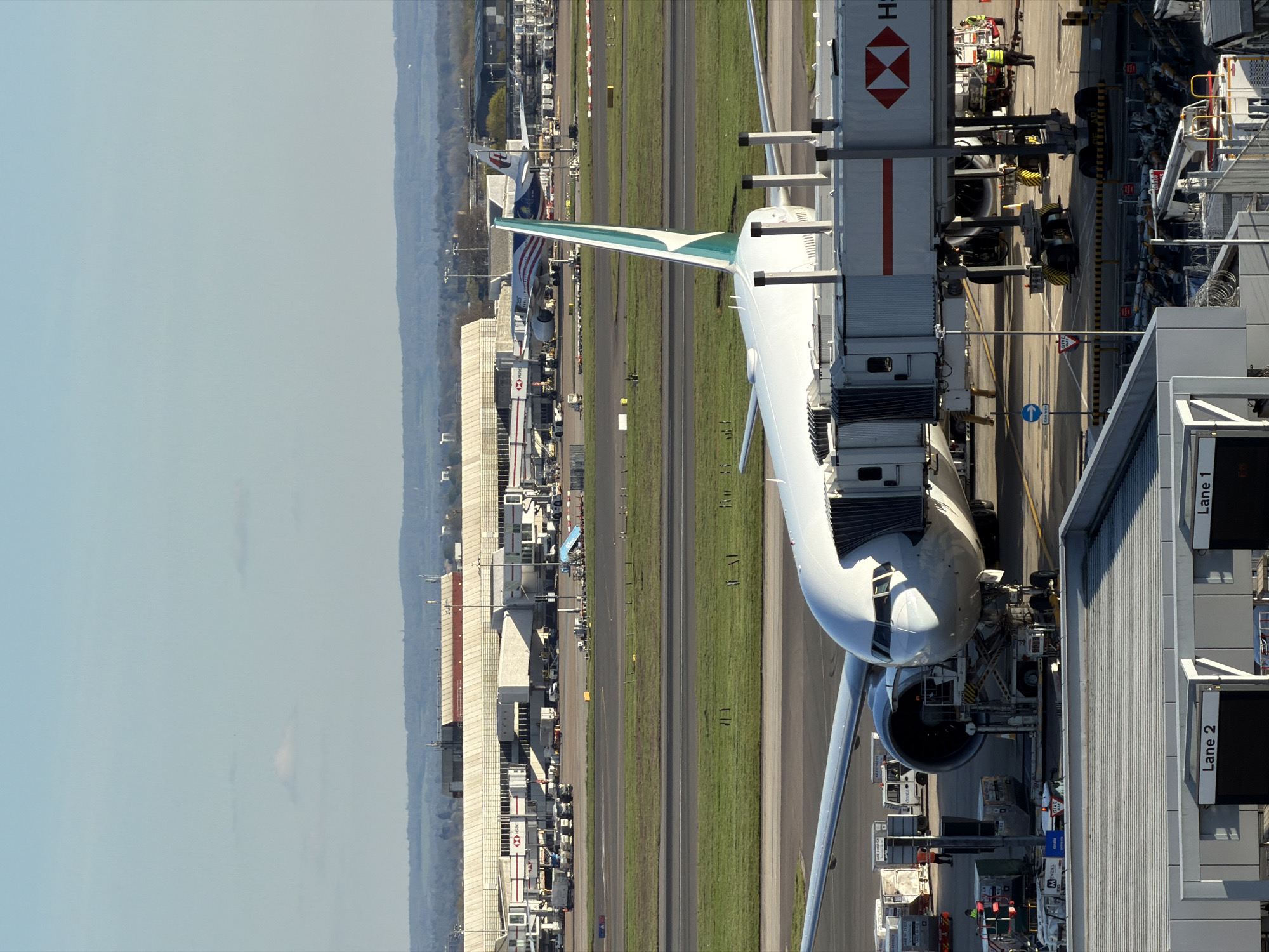 This image depicts an aerial view of an airport, likely in or near London, United Kingdom. A Cathay Pacific airplane is connected to a jet bridge at a gate. Various service vehicles and personnel are present around the aircraft. The airport features multiple runways and taxiways, along with adjacent buildings and infrastructure. In the distance, there are additional airplanes and airport facilities, with a hazy skyline visible. The image is taken from an elevated perspective, possibly from an airport control tower or a nearby building.