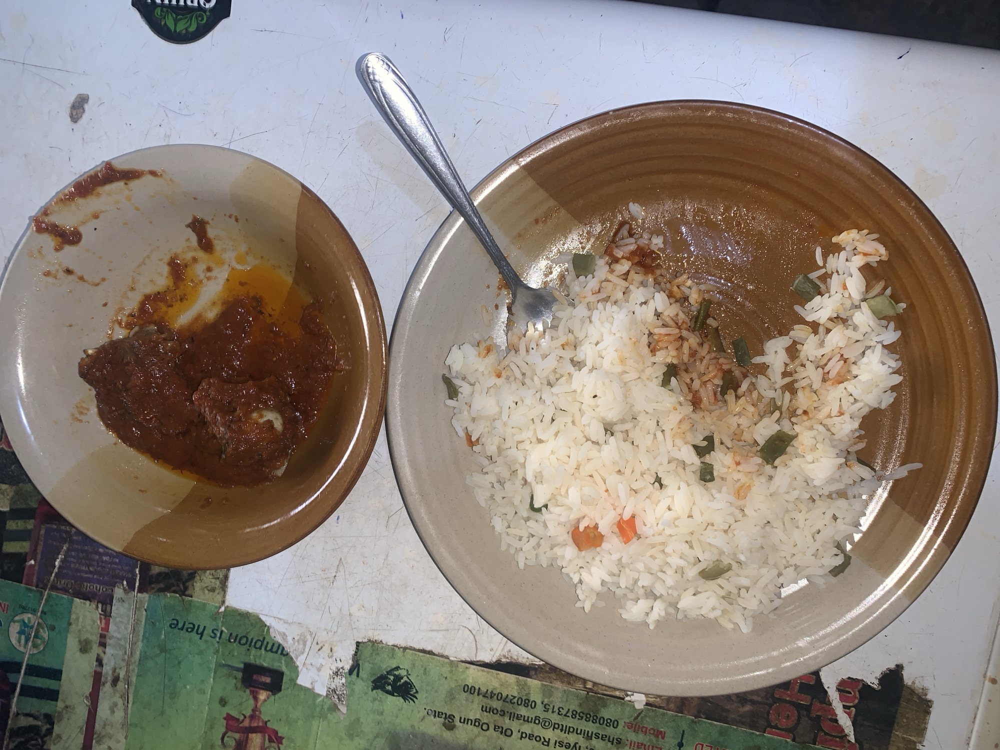 The image shows two bowls of food on a table. The bowl on the left contains a dark red sauce with chunks of meat. The bowl on the right contains white rice mixed with small pieces of green and orange vegetables. A silver fork rests on top of the rice. Parts of printed papers are visible beneath the bowls. One paper includes the text "Champion is here", "Iyesi Road, Ota, Ogun State," and a phone number and email address. Given the location information and the type of food, the scene is likely in Nigeria, though a more specific location like Kabong cannot be confirmed without more information.