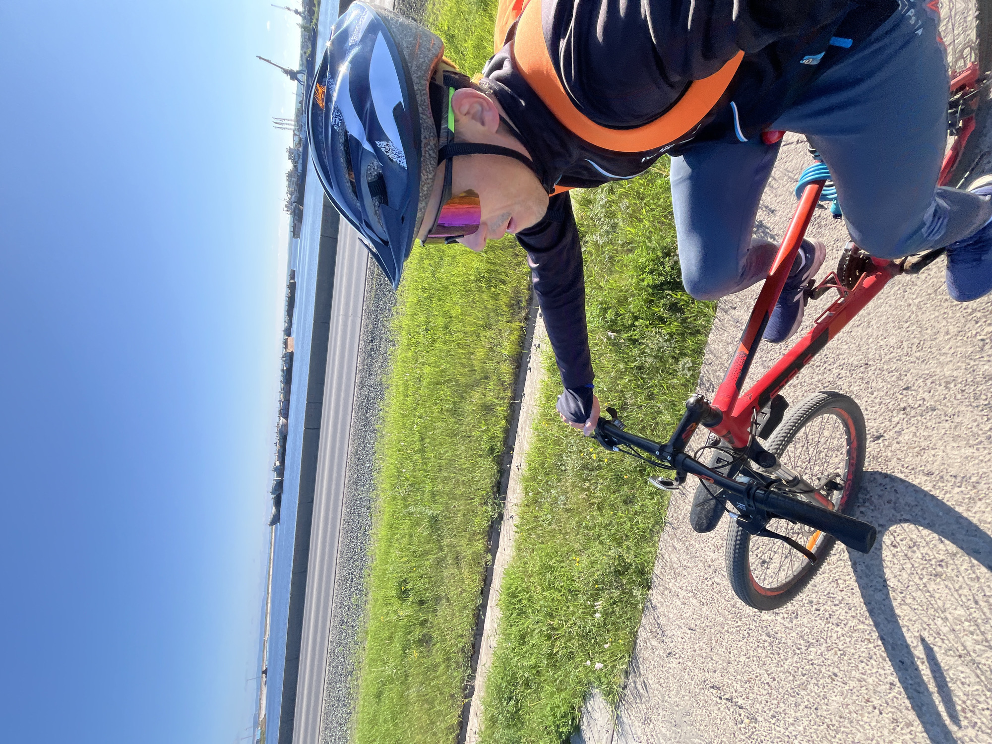 A man is riding a red bicycle on a sunny day. He is wearing a black helmet with white and blue accents, and reflective sunglasses with pink and orange lenses. He also has an orange backpack. The bicycle has a black handlebar and front wheel. The man is on a paved path next to a strip of green grass, with a road and a body of water with ships in the background. The sky is clear and blue. The scene appears to be in the city of Chaikovsky, Russia, likely near a port or industrial area, given the presence of ships and cranes in the distance. The sunlight casts a long shadow of the bicycle on the paved path.