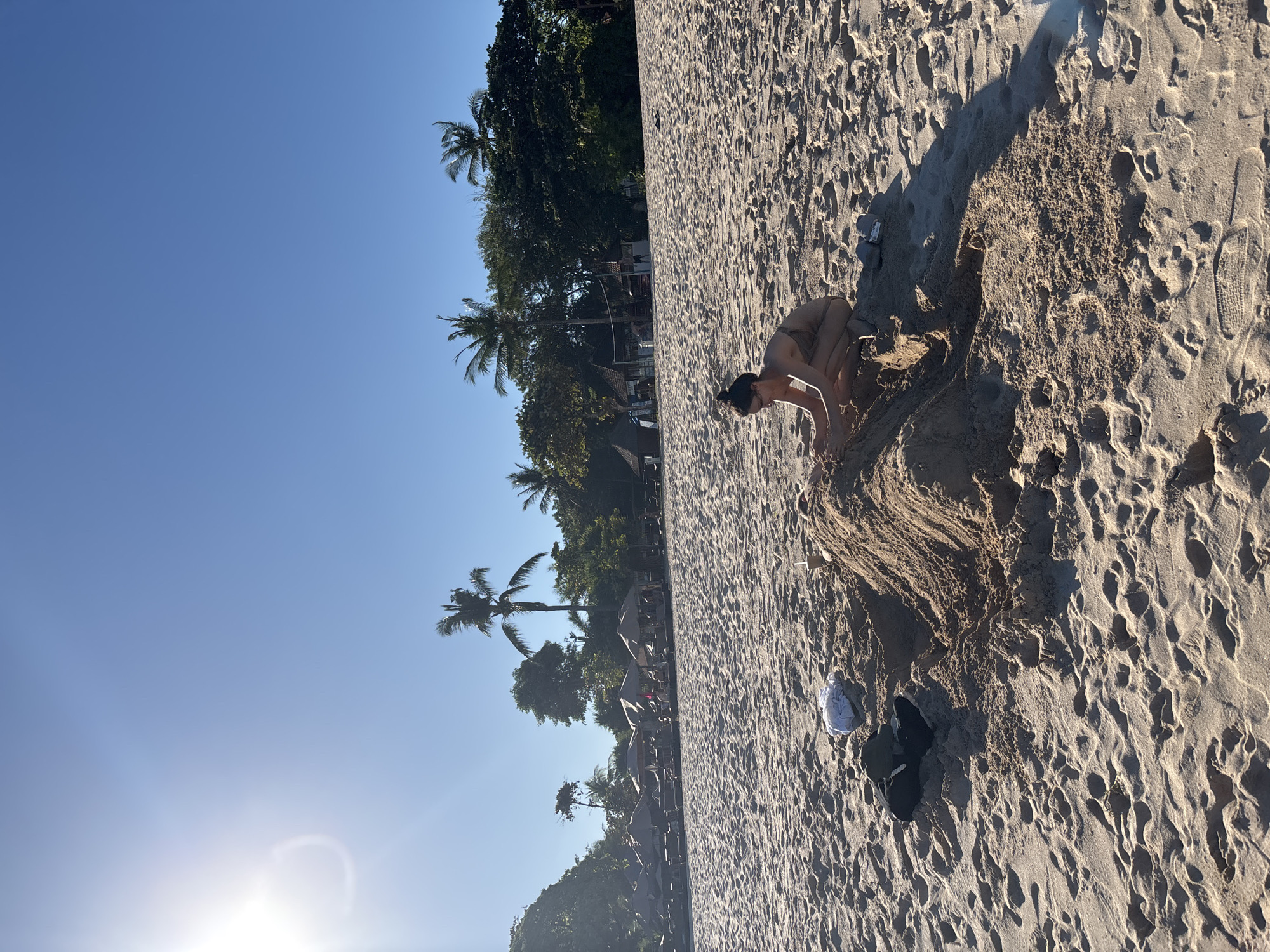 A person is playing in the sand on a beach in Kuta Selatan, Indonesia. The person is near a sand structure that is partially dug out in the middle. There are trees and a blue sky in the background. The sun is shining brightly. A white object sits next to the hole in the sand.