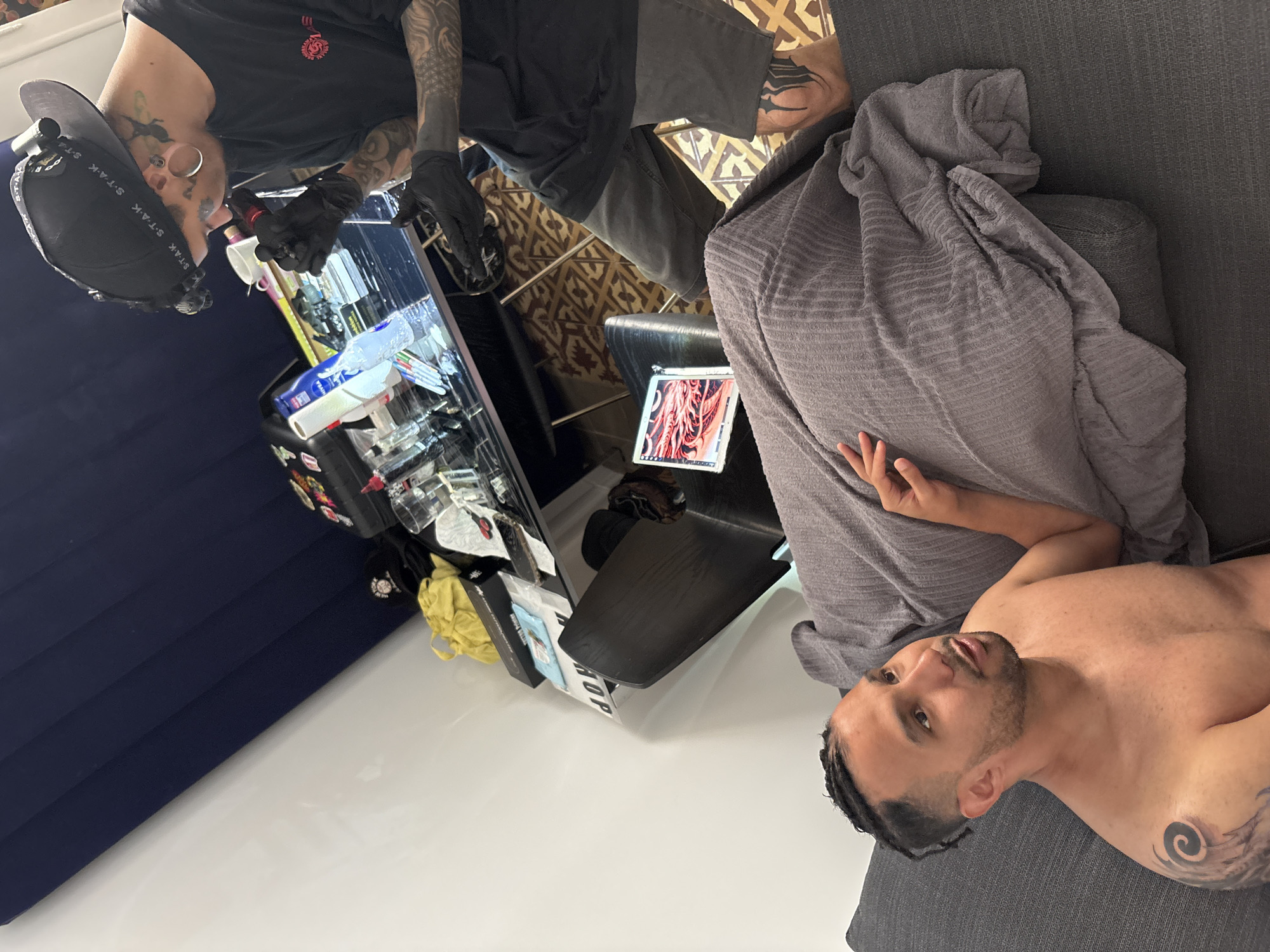 The image captures a scene where a tattoo artist is working on a client. The tattoo artist, wearing a black cap with the words "S-T-A-K" printed on it, is focused on their work, clad in a black t-shirt and gloves, their arms adorned with tattoos. They are positioned near a workstation equipped with tattoo equipment and supplies, placed on a stand that has the word "AOP" written on it.
The client, lying on a grey upholstered sofa covered with a blanket, is receiving the tattoo. Part of their upper torso is exposed, revealing an existing tattoo on their shoulder. They are looking up towards the artist. A digital display showing tattoo designs is placed near the sofa on a small black table.
The location appears to be an indoor setting, likely a tattoo studio. Given that this model is not trained to identify locations, there is no way to infer the location in the city of Barcelona, Spain. The environment seems well-lit, and the presence of the tattoo equipment indicates the professional nature of the setting.